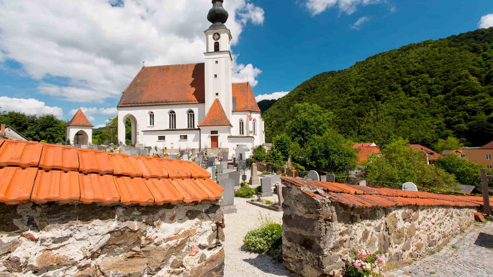 A picturesque view of a traditional white church with a red-tiled roof and a prominent bell tower in the town of Engelhartszell, Austria, with a cemetery and green hills behind it.