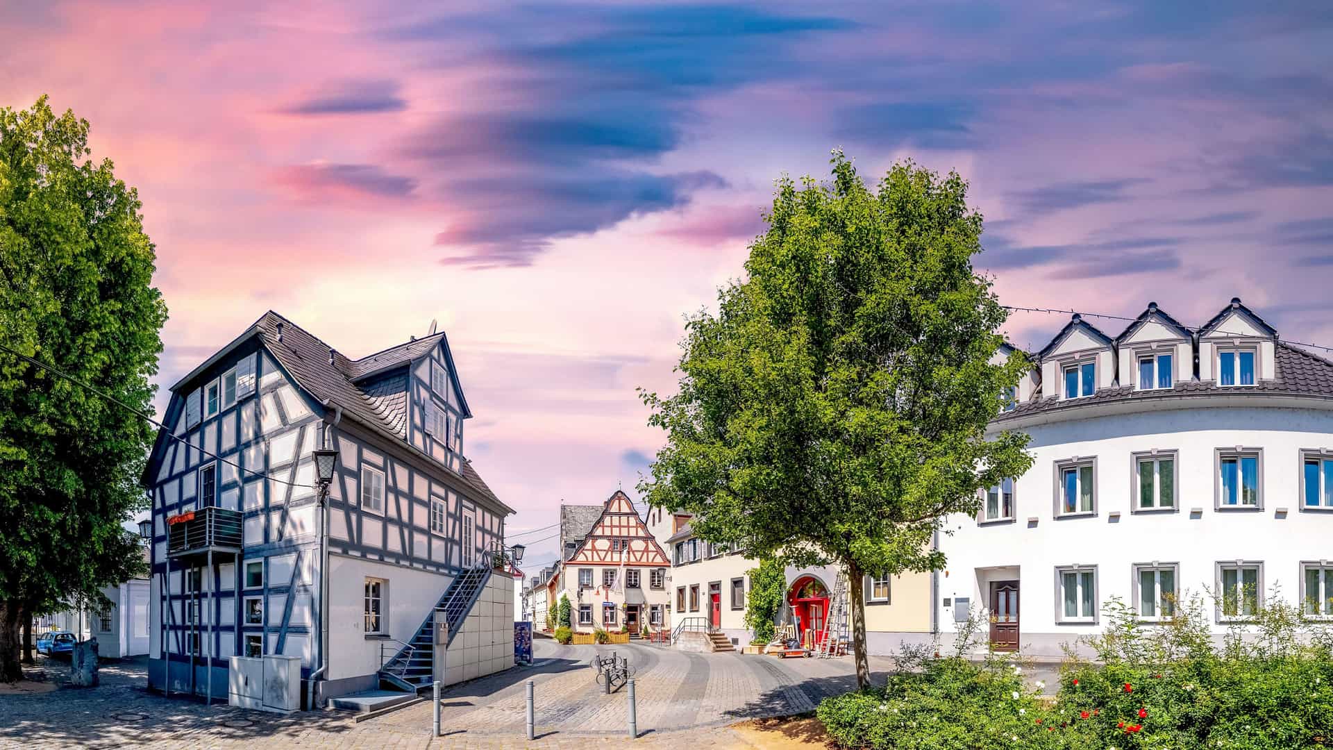 A charming panoramic shot of the old town square in Engers, Germany, featuring historic half-timbered houses and a large green tree under a dramatic, colorful sunset sky.