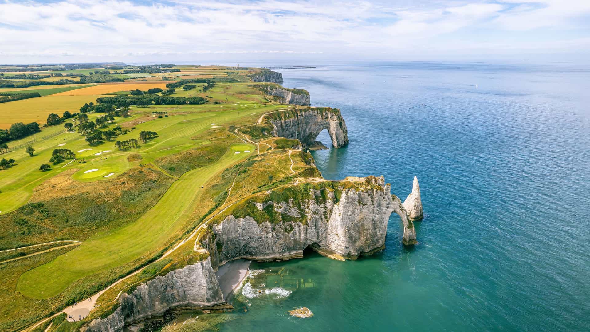 An aerial view of the iconic white chalk cliffs of Étretat, Normandy, with natural arches and a sea stack, surrounded by lush green fields and the expansive English Channel.