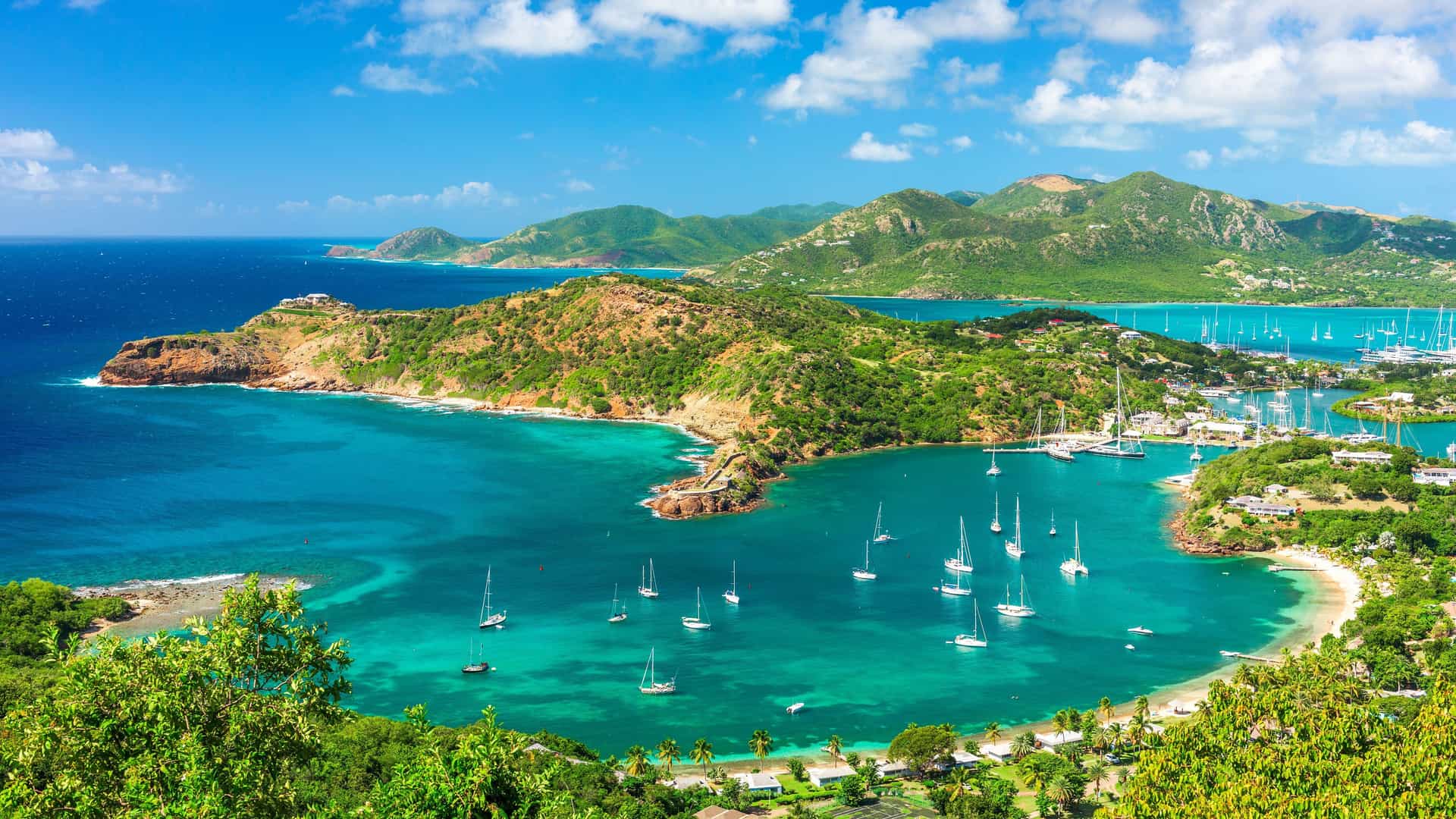 A breathtaking aerial shot of English Harbour in Antigua, with a pristine turquoise bay filled with sailboats, surrounded by lush green hills and a stunning coastline.