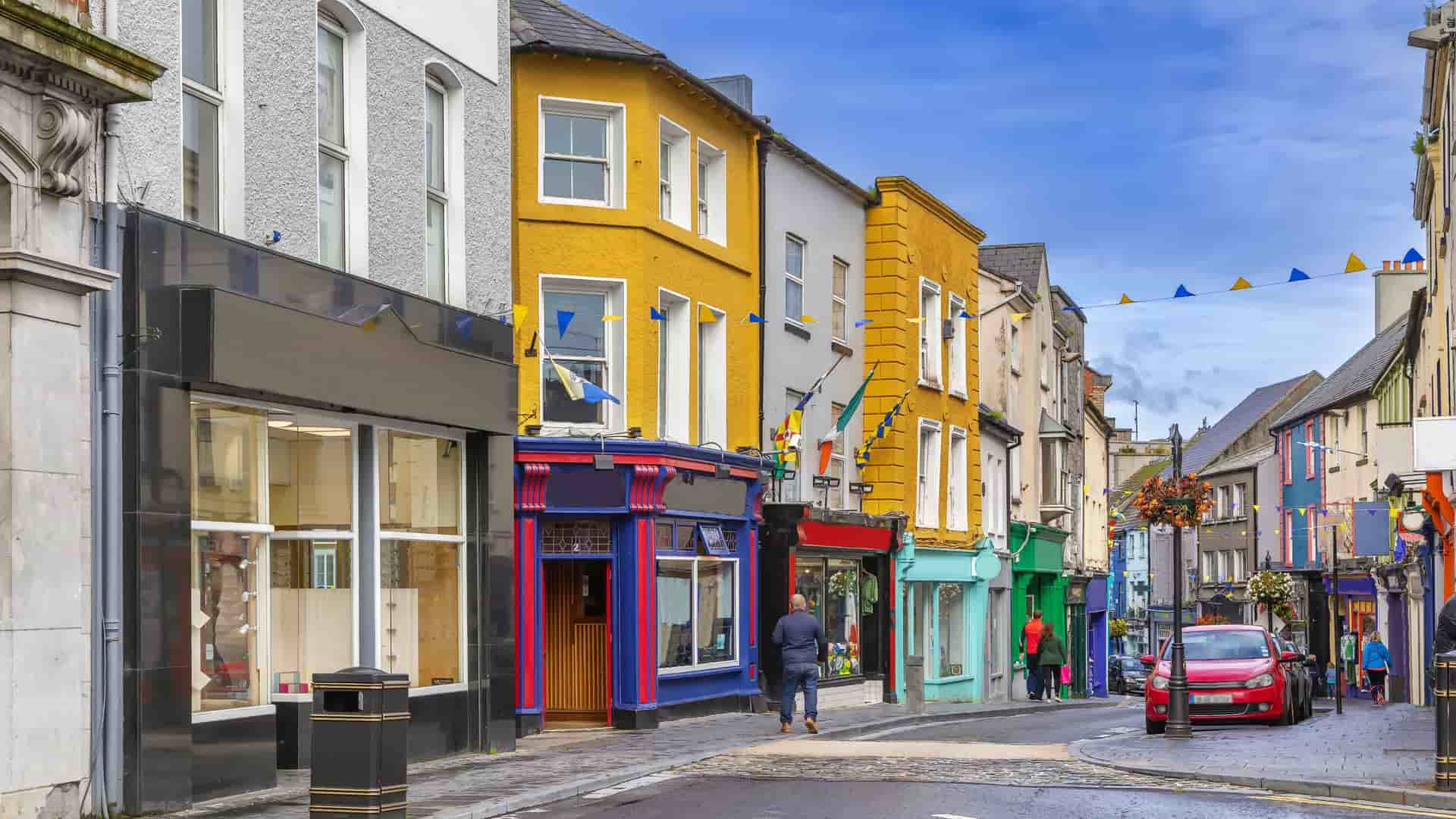 "A charming street view in the town of Ennis, Ireland, with colorful storefronts and traditional pubs lining the cobblestone street under a partially cloudy sky. "