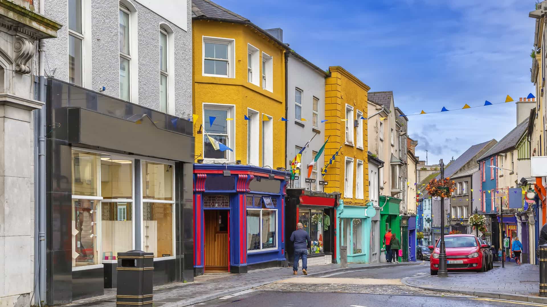 "A charming street view in the town of Ennis, Ireland, with colorful storefronts and traditional pubs lining the cobblestone street under a partially cloudy sky. "