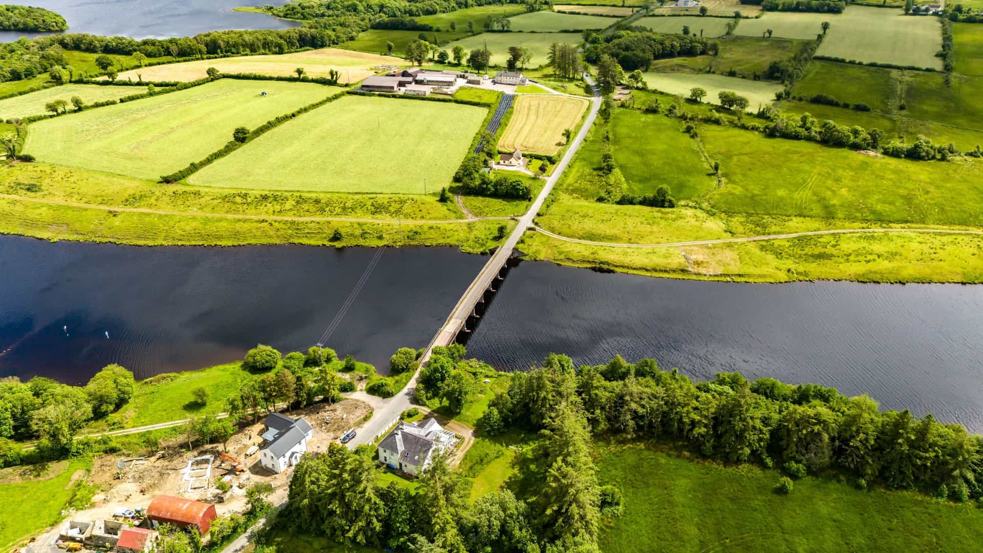 Aerial view of a bridge crossing a dark, flowing river, surrounded by a patchwork of vibrant green farmlands and scattered houses near Enniskillen, Northern Ireland.