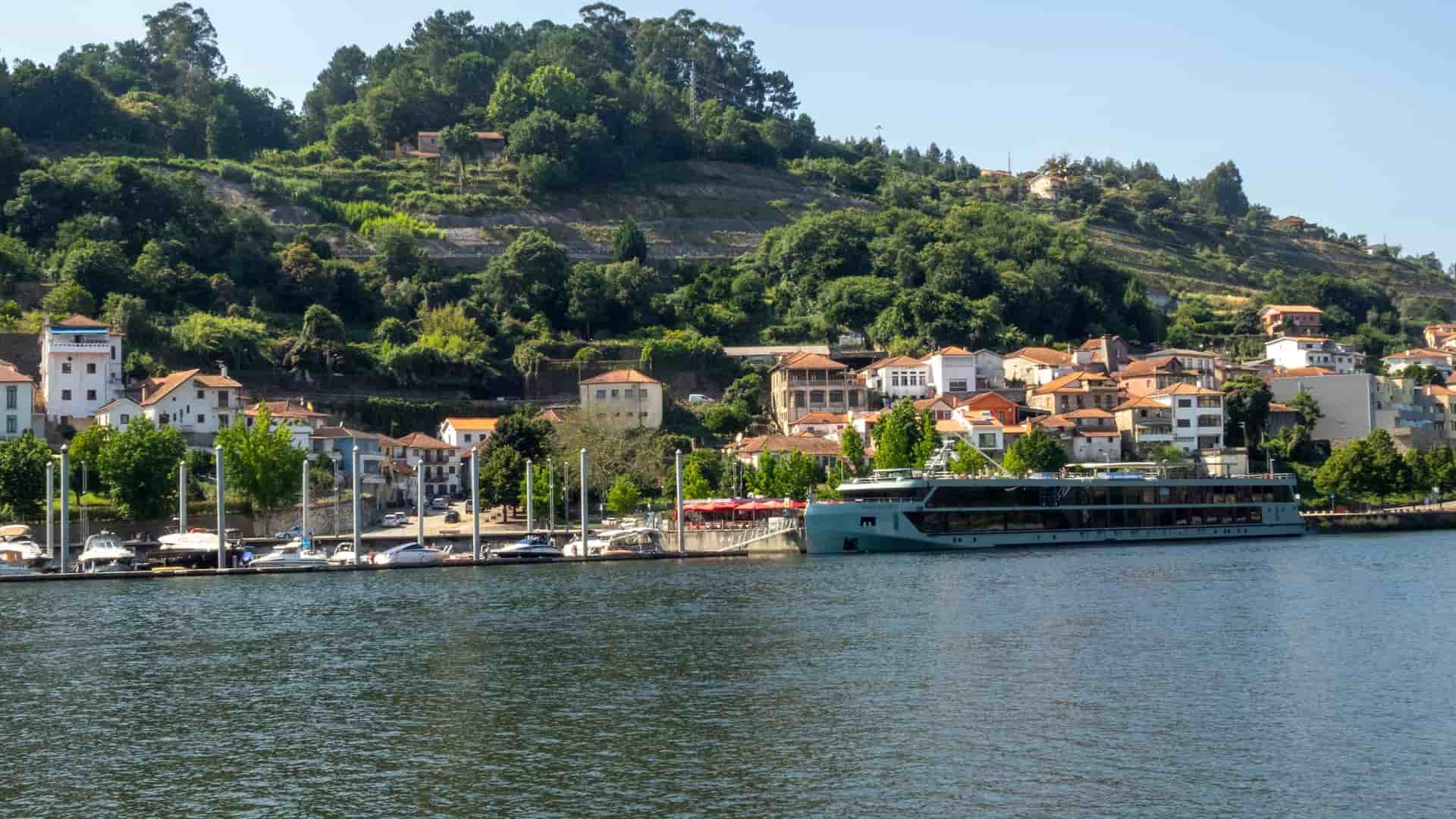 A scenic view of Entre-os-Rios, Portugal, with a large river cruise ship docked at a waterfront marina, and historic buildings nestled on a steep, tree-covered hillside in the background.