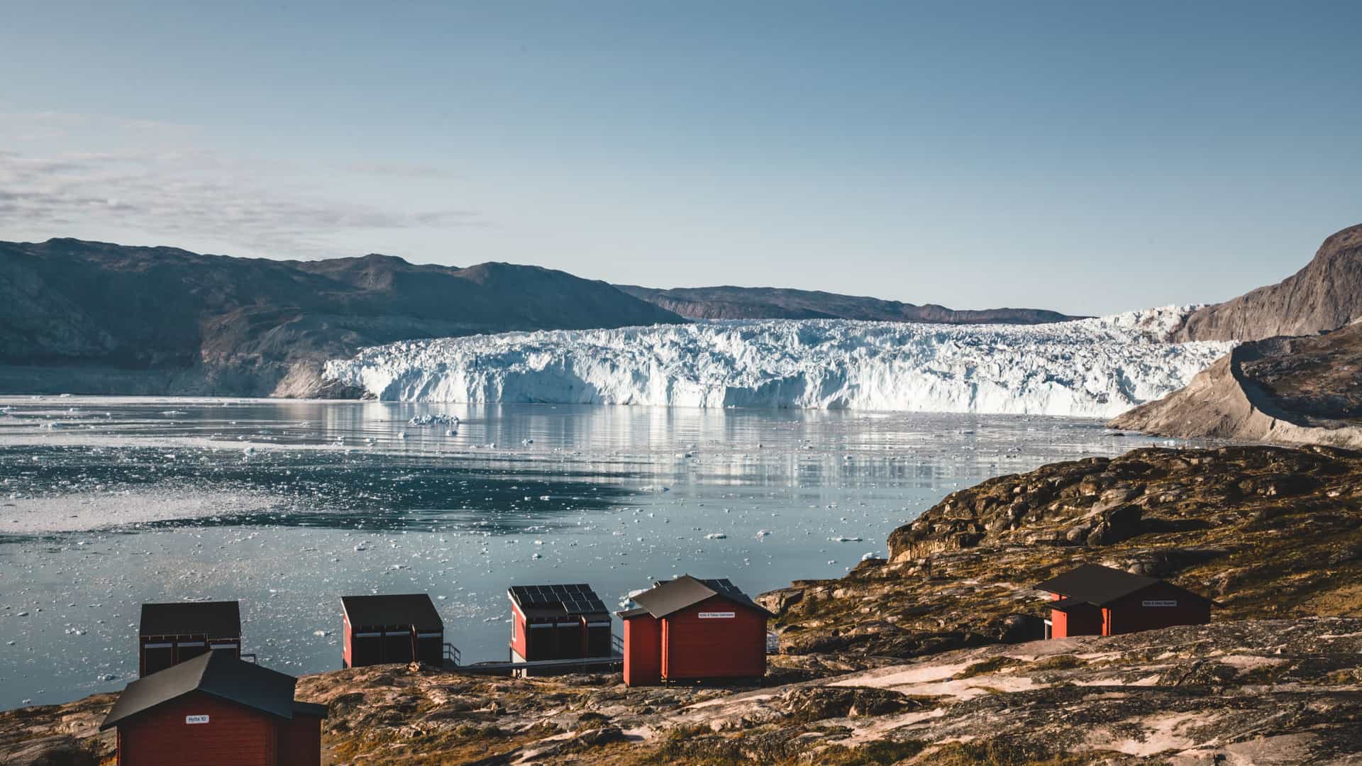 A panoramic view of the massive Eqip Sermia Glacier in Greenland, with a cluster of red huts on the rocky shore and ice chunks floating in the water under a clear blue sky.