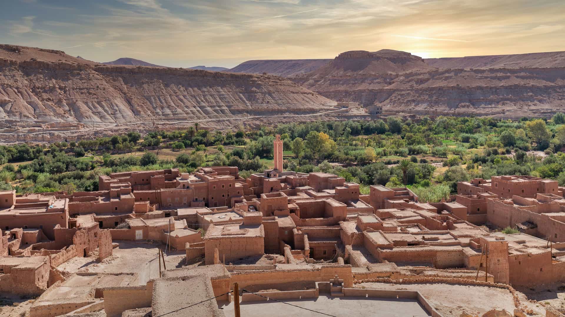 "A high-angle view of a traditional clay village in Erfoud, Morocco, with a single minaret in the center and a lush green oasis of date palms and other trees in a valley, surrounded by the barren, rocky landscape of the Atlas Mountains. "