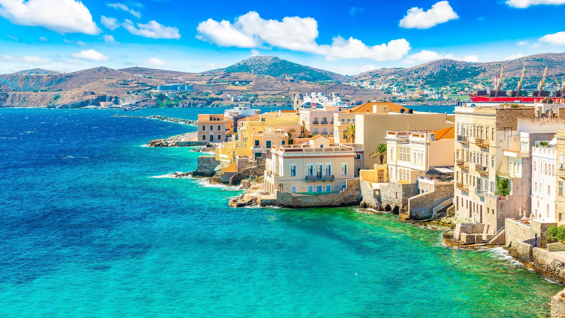 A breathtaking view of Ermoupolis on the island of Syros, Greece, showcasing beautiful neoclassical buildings perched on the rocky coastline, with vibrant turquoise water and rolling hills in the background.