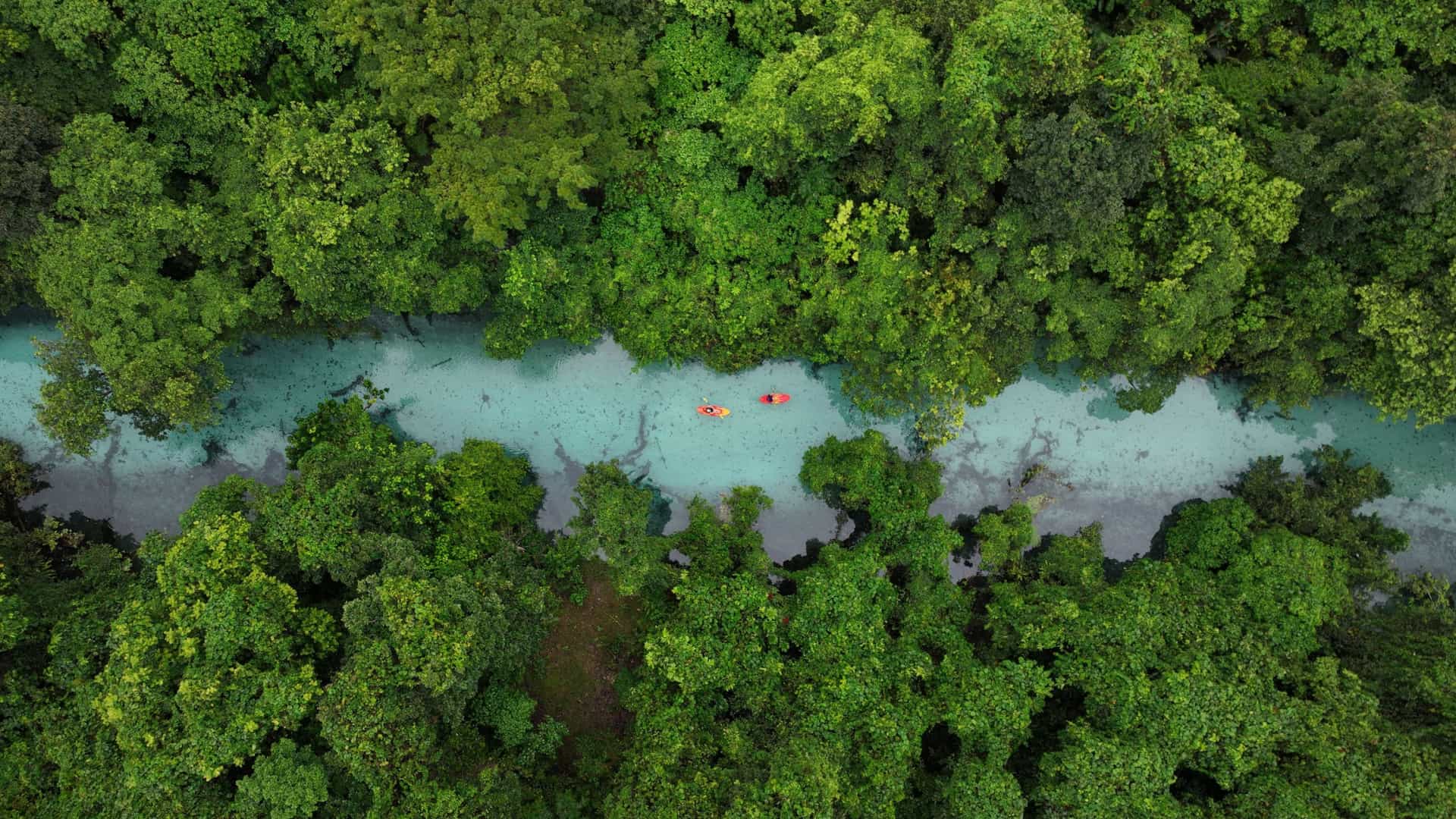 An aerial view of two red kayaks floating on a stunning, clear turquoise river surrounded by dense, lush green rainforest on Espiritu Santo Island, Vanuatu.