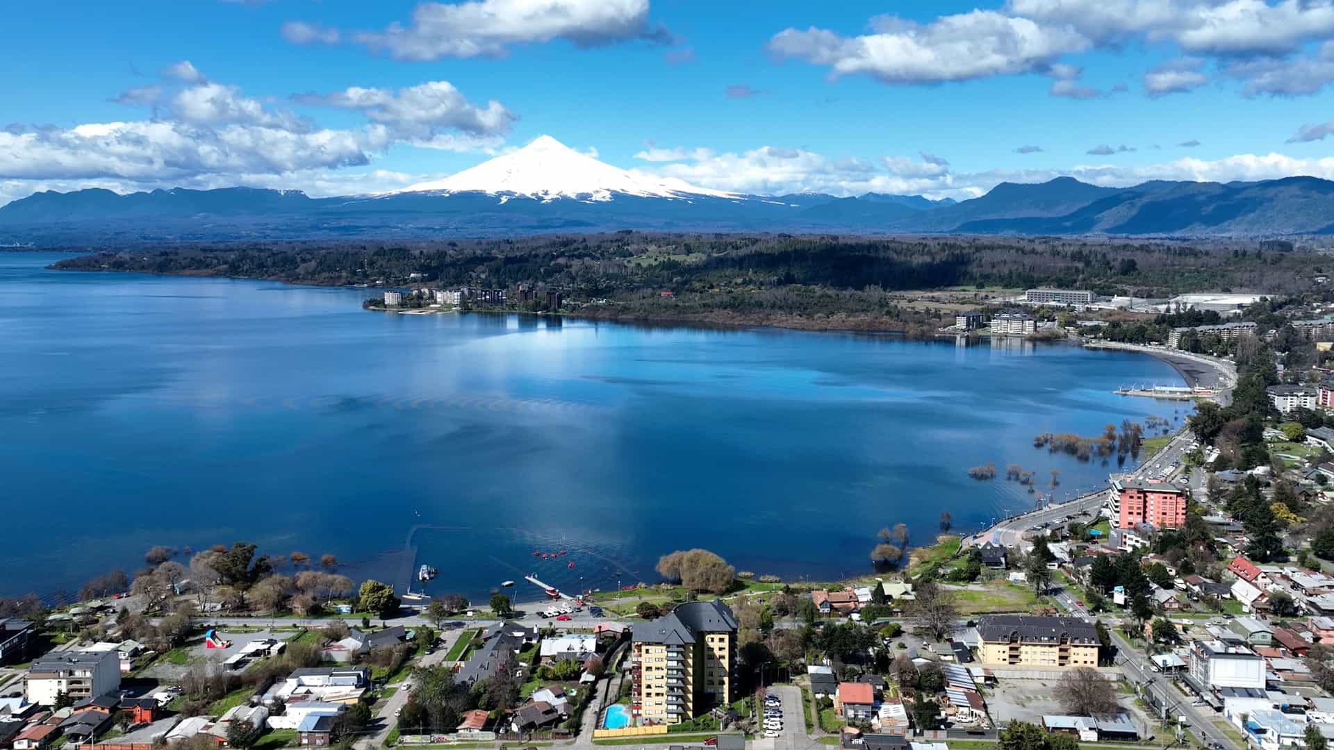 An aerial view of a lake and a lakeside town with the Villarrica Volcano, a snow-capped mountain, prominently featured in the background under a blue sky with scattered clouds.