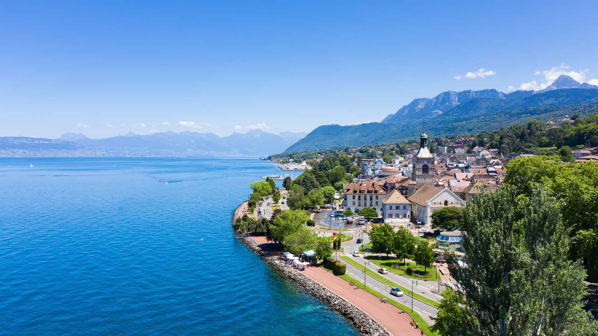 An aerial view of the picturesque town of Évian-les-Bains on the shore of Lake Geneva, with a scenic promenade, historic buildings, and the French Alps in the background.