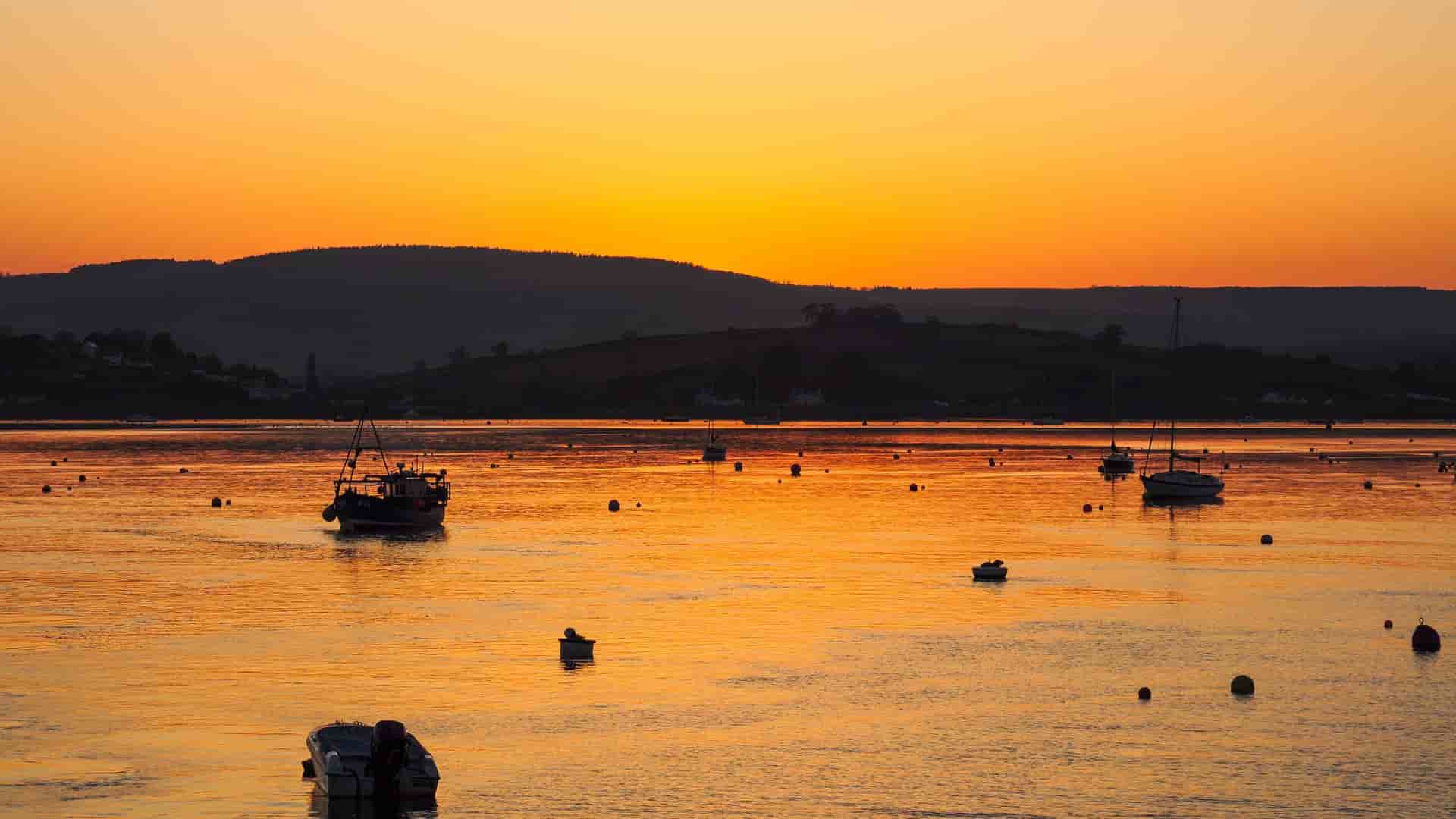A tranquil sunset view over the Exe Estuary in Exmouth, Devon, with fishing boats and sailboats dotting the calm, reflective orange water and hills silhouetted in the background.