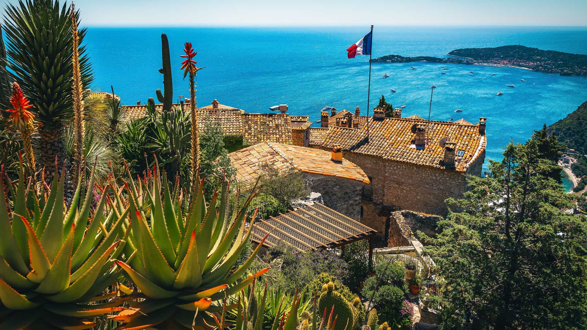 A beautiful landscape shot of the medieval village of Èze, France, overlooking the azure waters of the French Riviera, with a variety of lush, exotic plants in the foreground.