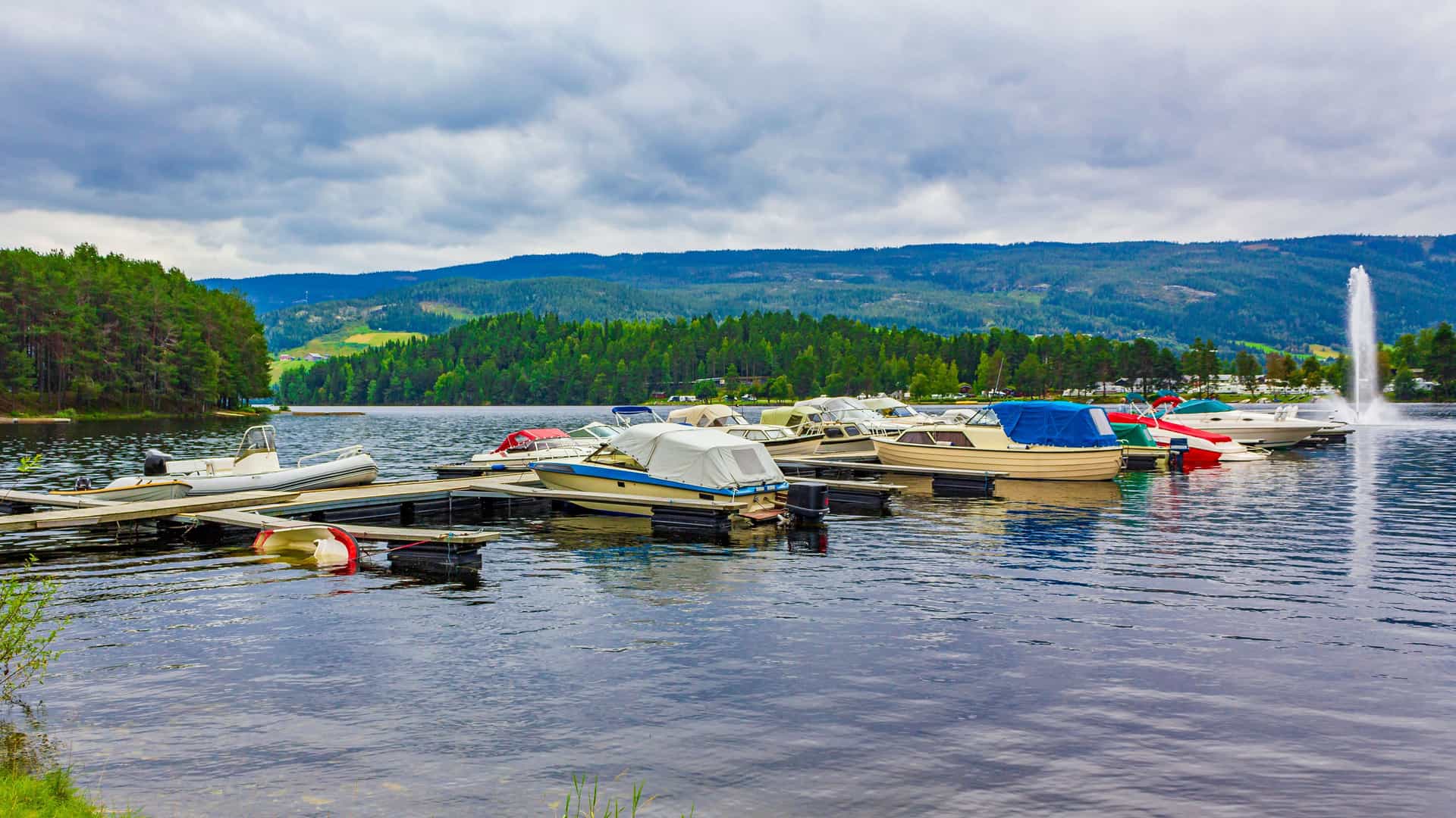 "A small flotilla of motorboats and personal watercraft are tied to a floating dock on a tranquil lake with a fountain jetting water high into the air, surrounded by a lush green pine forest in Fagernes, Norway. "