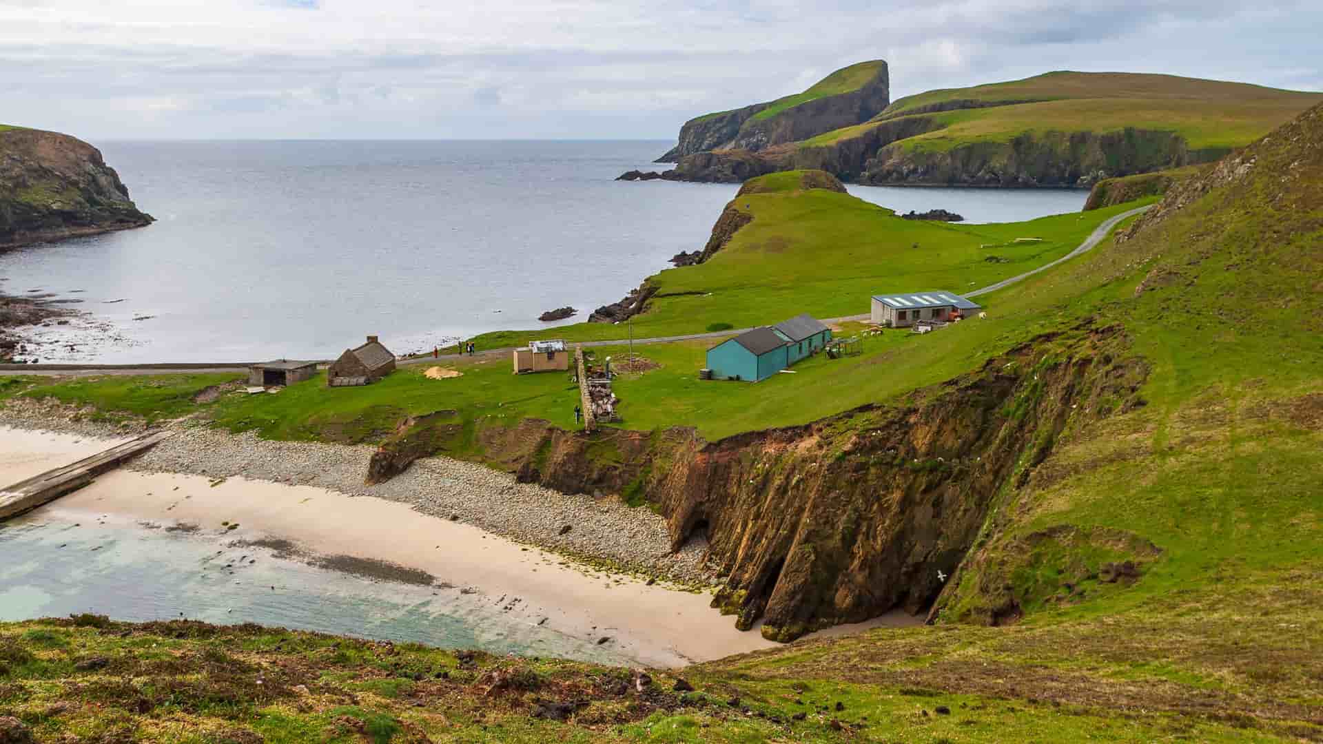 A panoramic view of a small bay and sandy beach on Fair Isle, a remote island in the Shetlands, Scotland, with a few buildings, a winding road, and dramatic green cliffs.