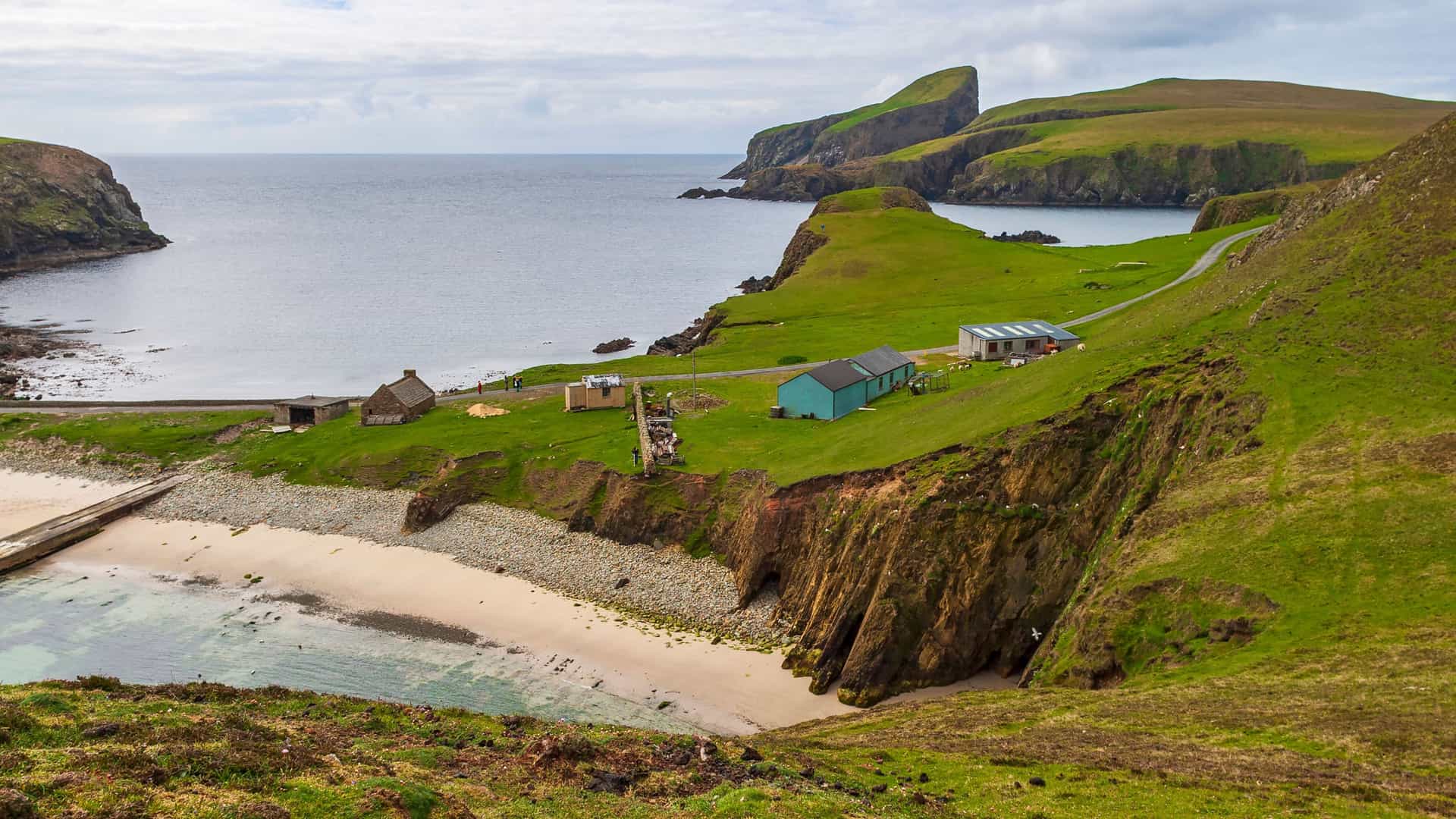 A panoramic view of a small bay and sandy beach on Fair Isle, a remote island in the Shetlands, Scotland, with a few buildings, a winding road, and dramatic green cliffs.