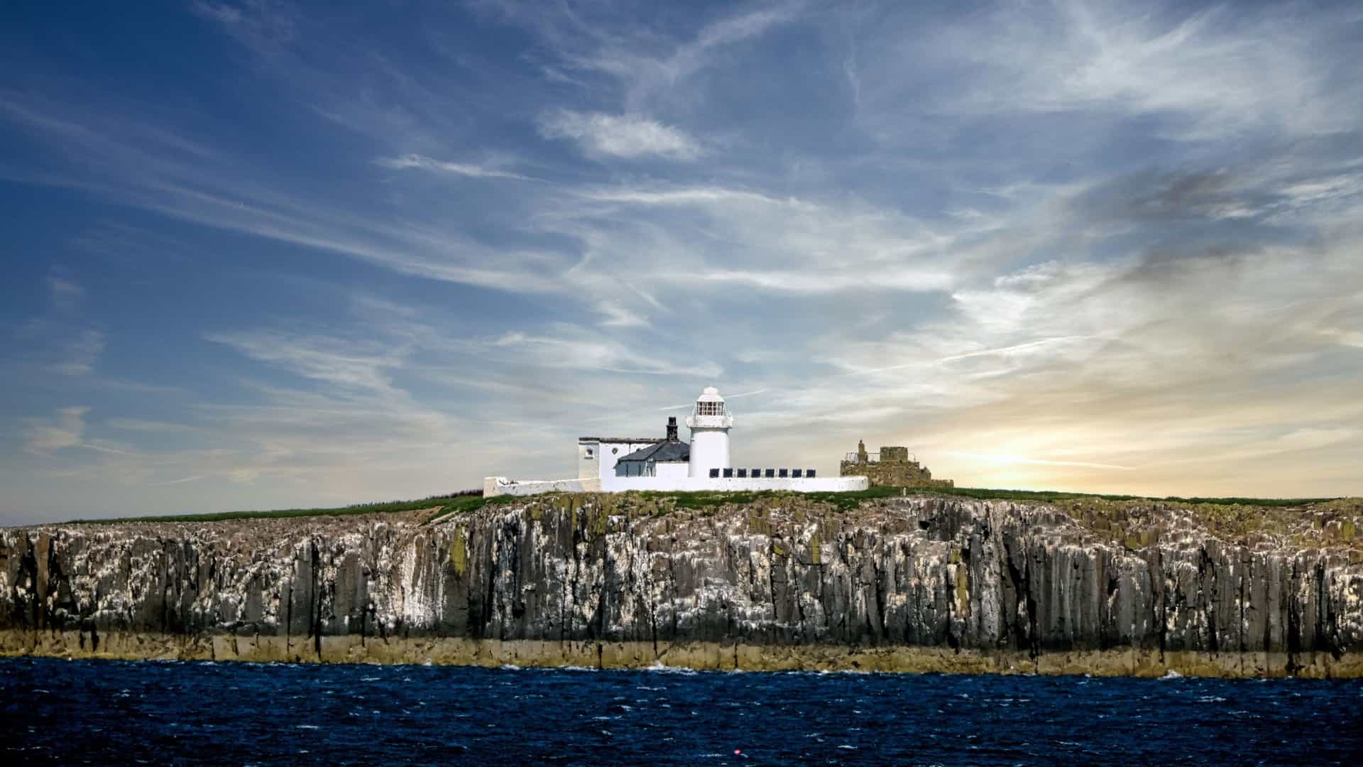 A lighthouse and a cottage are perched on the rocky cliffs of the Farne Islands, a significant breeding ground for seabirds, with a dramatic, cloudy sky and rough seas in the foreground.