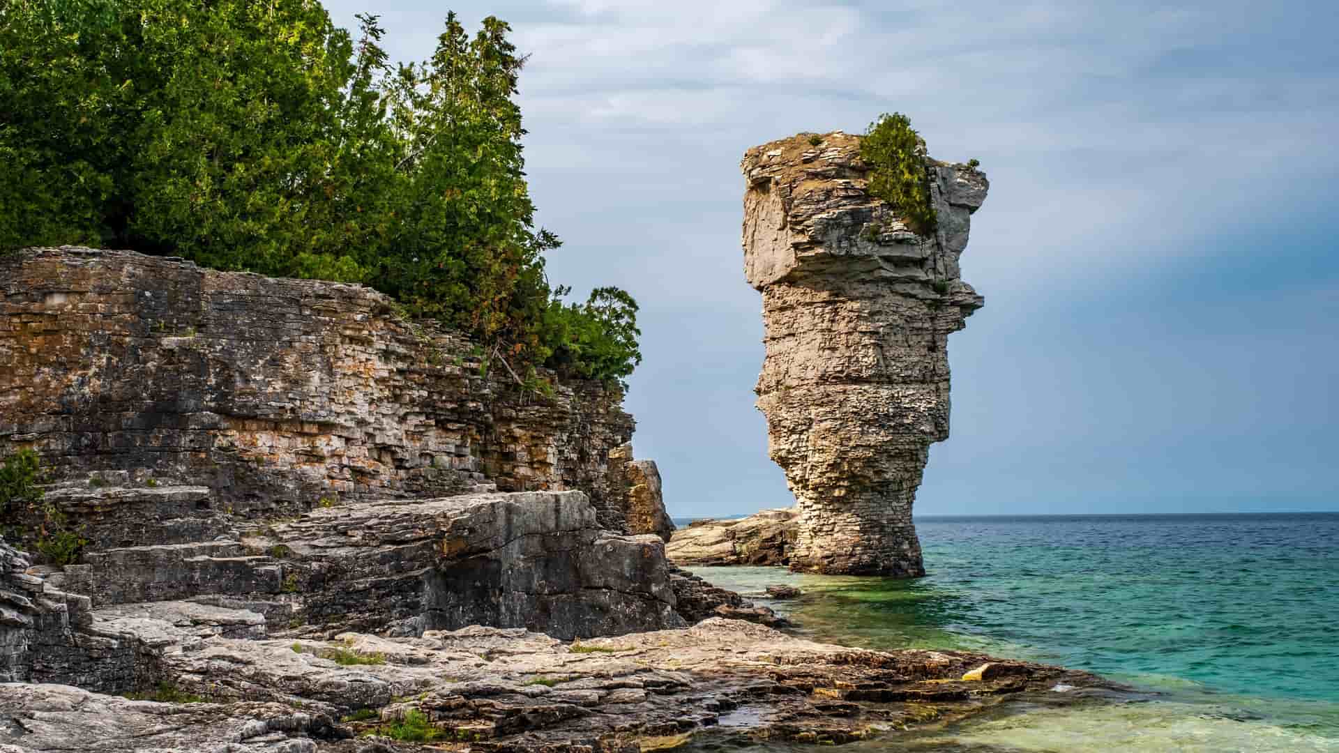 A striking landscape image of the "Flowerpot" sea stack, a unique rock formation in Fathom Five National Marine Park, surrounded by clear turquoise water and rocky cliffs topped with green trees.