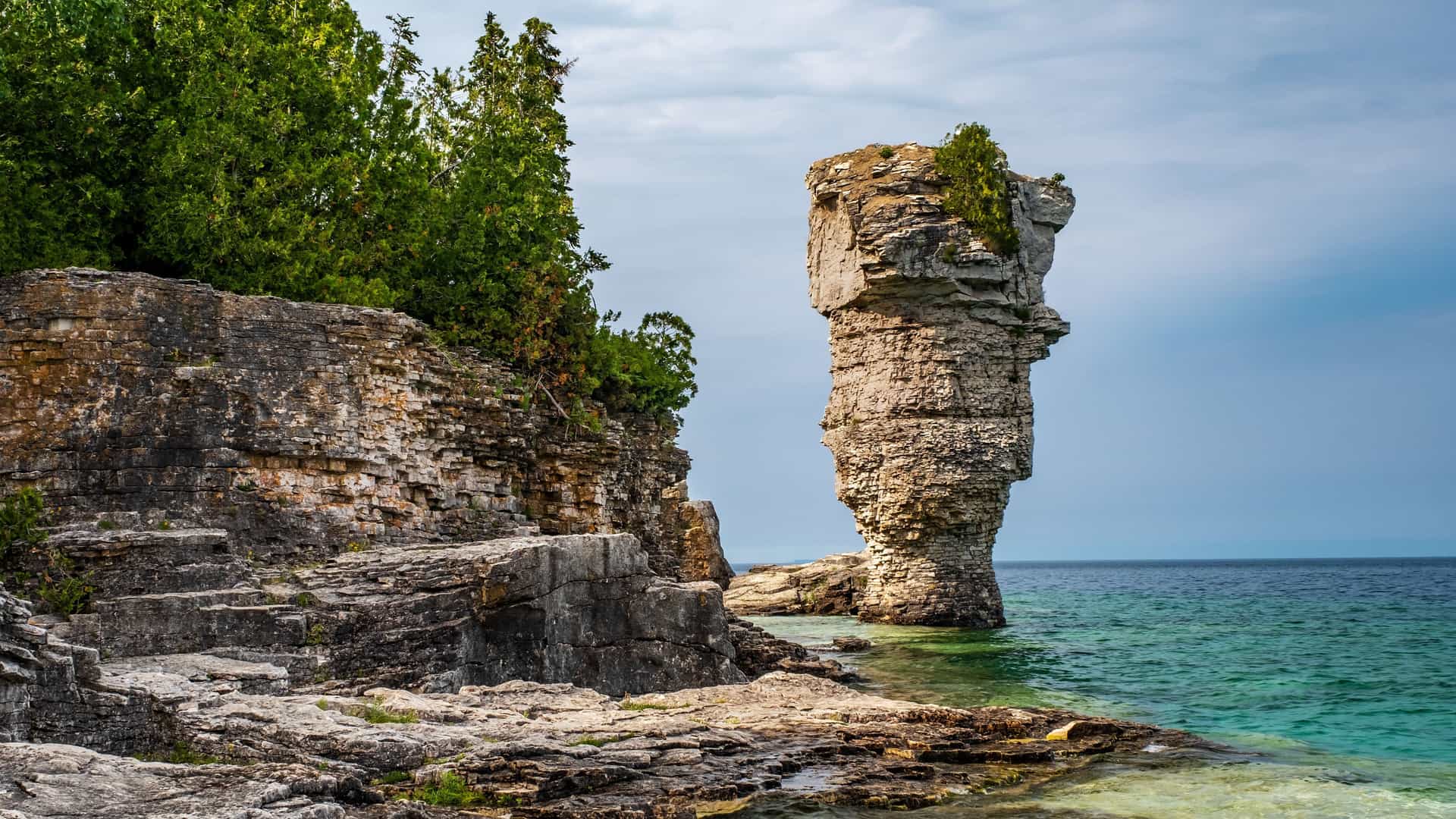 A striking landscape image of the "Flowerpot" sea stack, a unique rock formation in Fathom Five National Marine Park, surrounded by clear turquoise water and rocky cliffs topped with green trees.