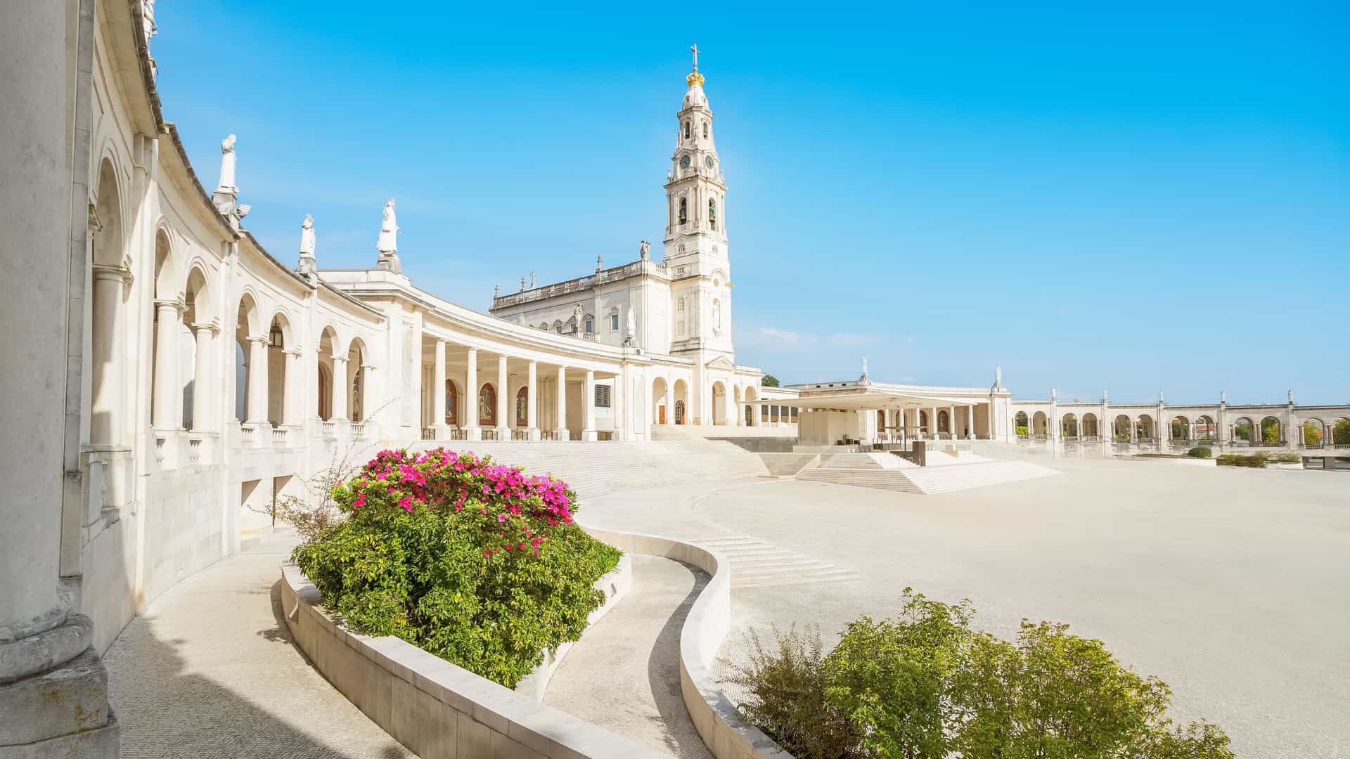 "A panoramic view of the Sanctuary of Fátima in Portugal, a vast, open plaza surrounded by a curving colonnade with a large church and a prominent bell tower. "