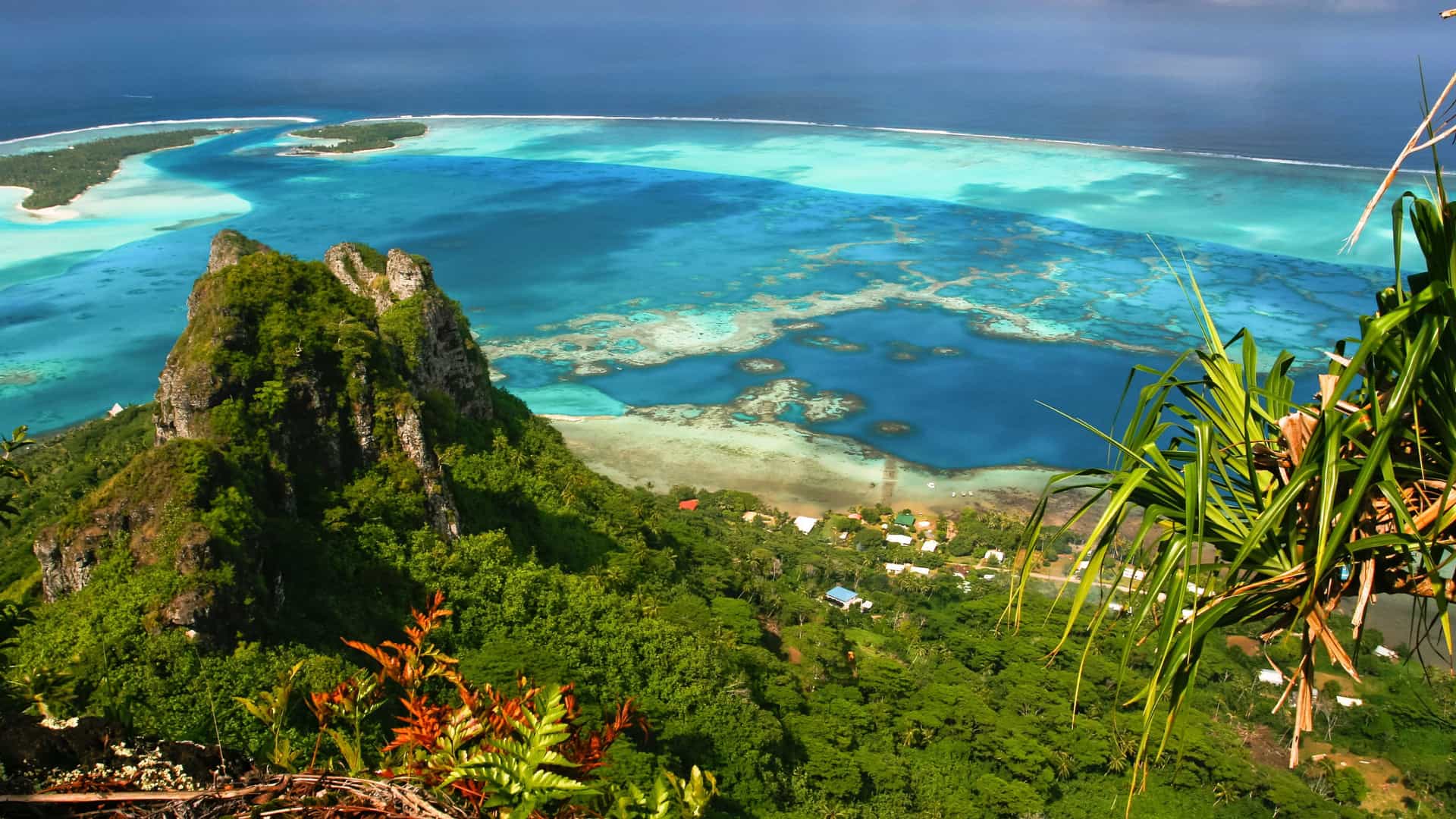 A dramatic aerial view overlooking the lush, mountainous landscape of Fatu Hiva, one of the Marquesas Islands, with a stunning turquoise lagoon and the deep blue Pacific Ocean in the background.