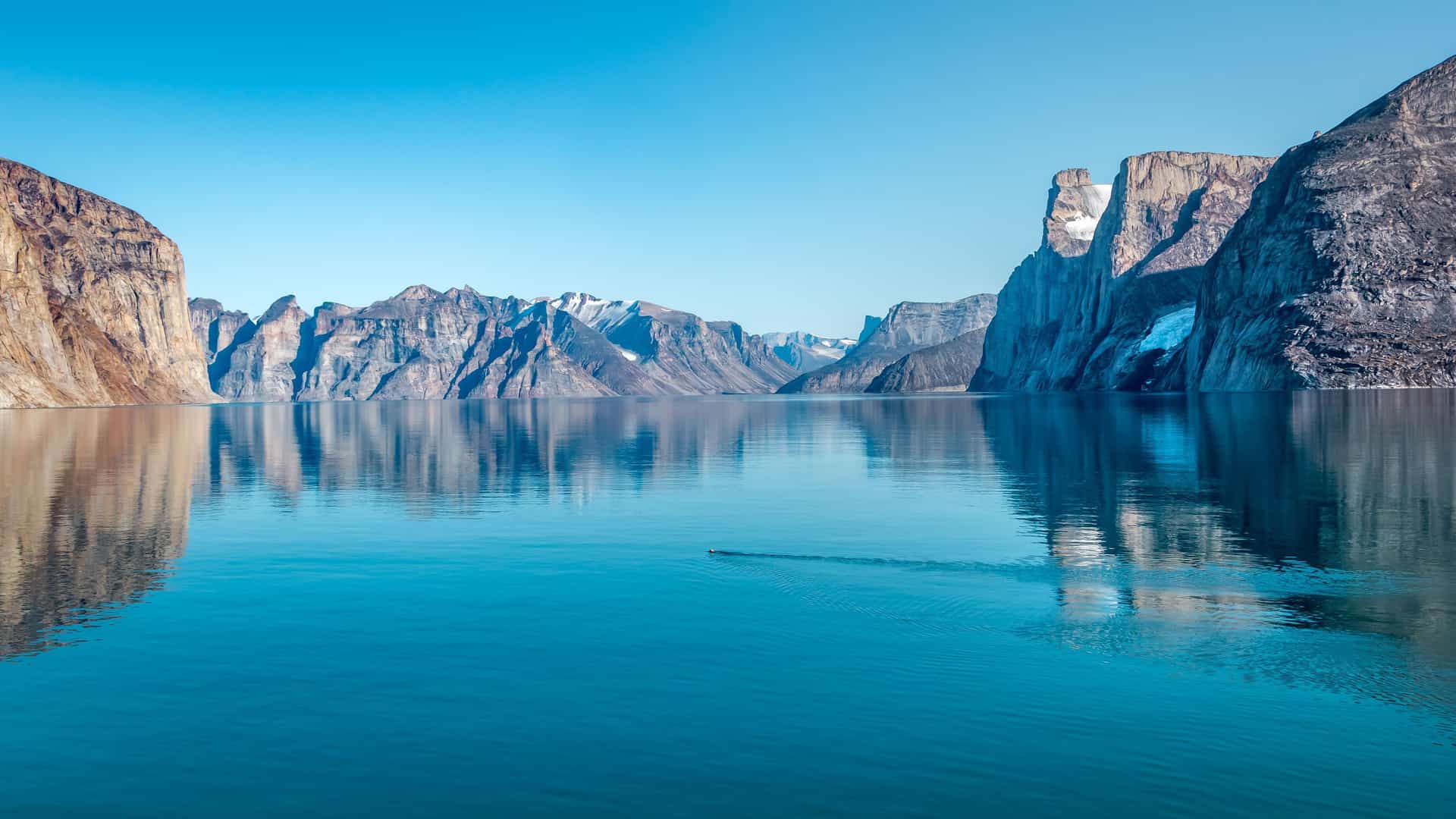 A breathtaking view of Fecham Bay, with towering, sheer cliffs and mountains on both sides reflected perfectly in the still, glassy water under a clear blue sky.