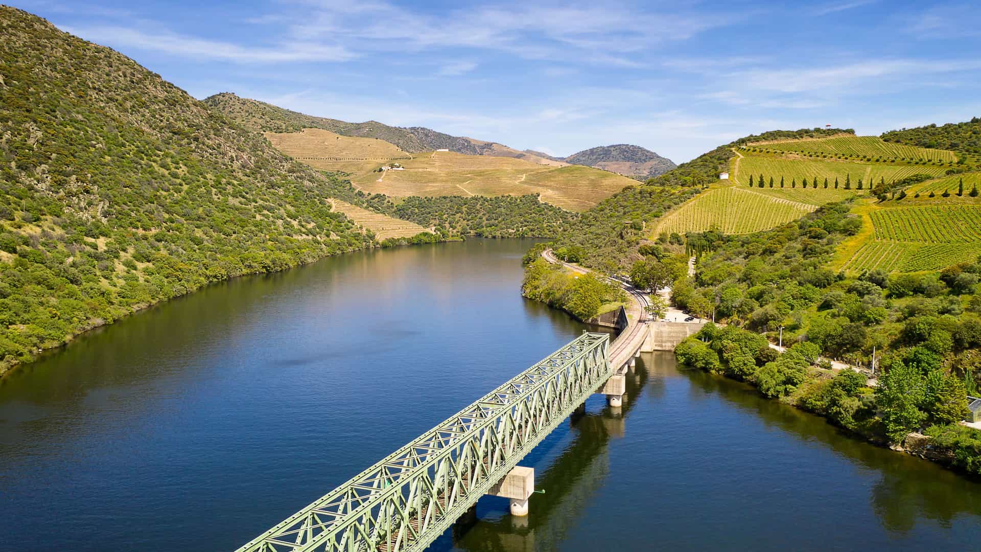 An aerial view of a green railway bridge crossing the tranquil Douro River, surrounded by the steep, terraced vineyards and lush hills of Ferradosa in Portugal.