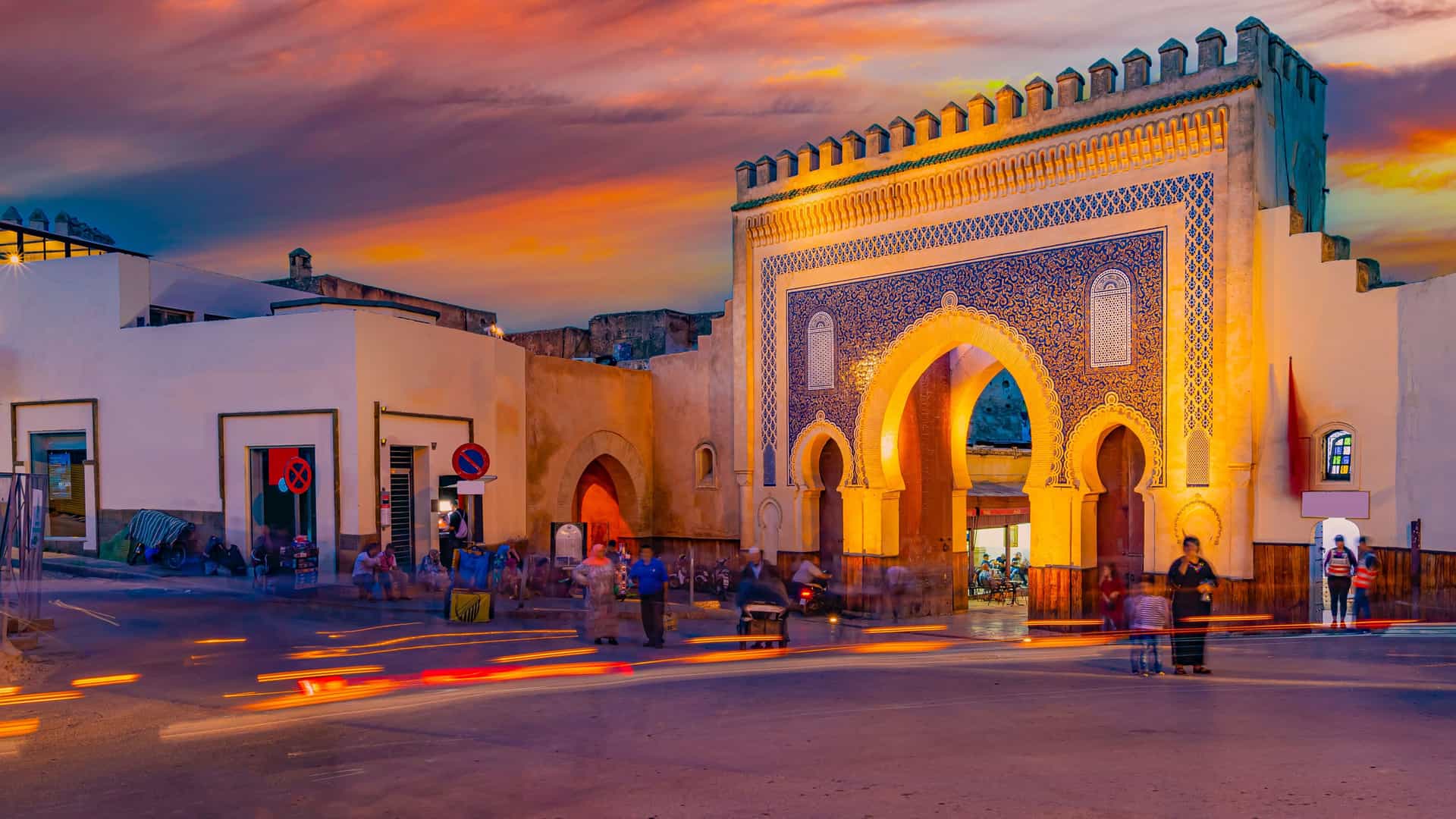 "A beautiful sunset view of the iconic Bab Bou Jeloud gate in Fez, Morocco, with its intricate blue and green tile mosaics, glowing in the light as people bustle through the archway. "