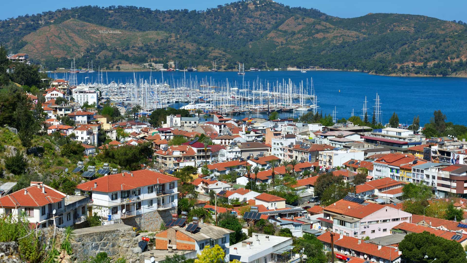 An elevated view of the beautiful coastal town of Fethiye, Turkey, showing a bustling marina filled with countless sailboats and a harbor surrounded by houses with red roofs and green hills.