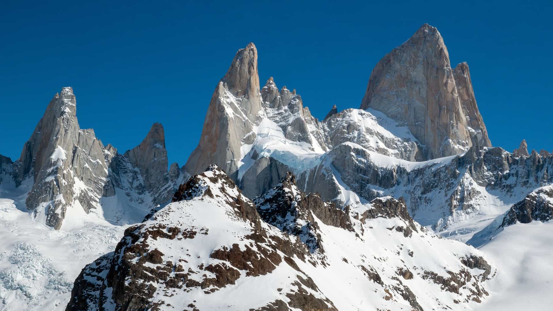 Jagged, snow-covered, light-colored granite peaks rise dramatically against a clear blue sky in Fiordo de las Montañas, Chile.