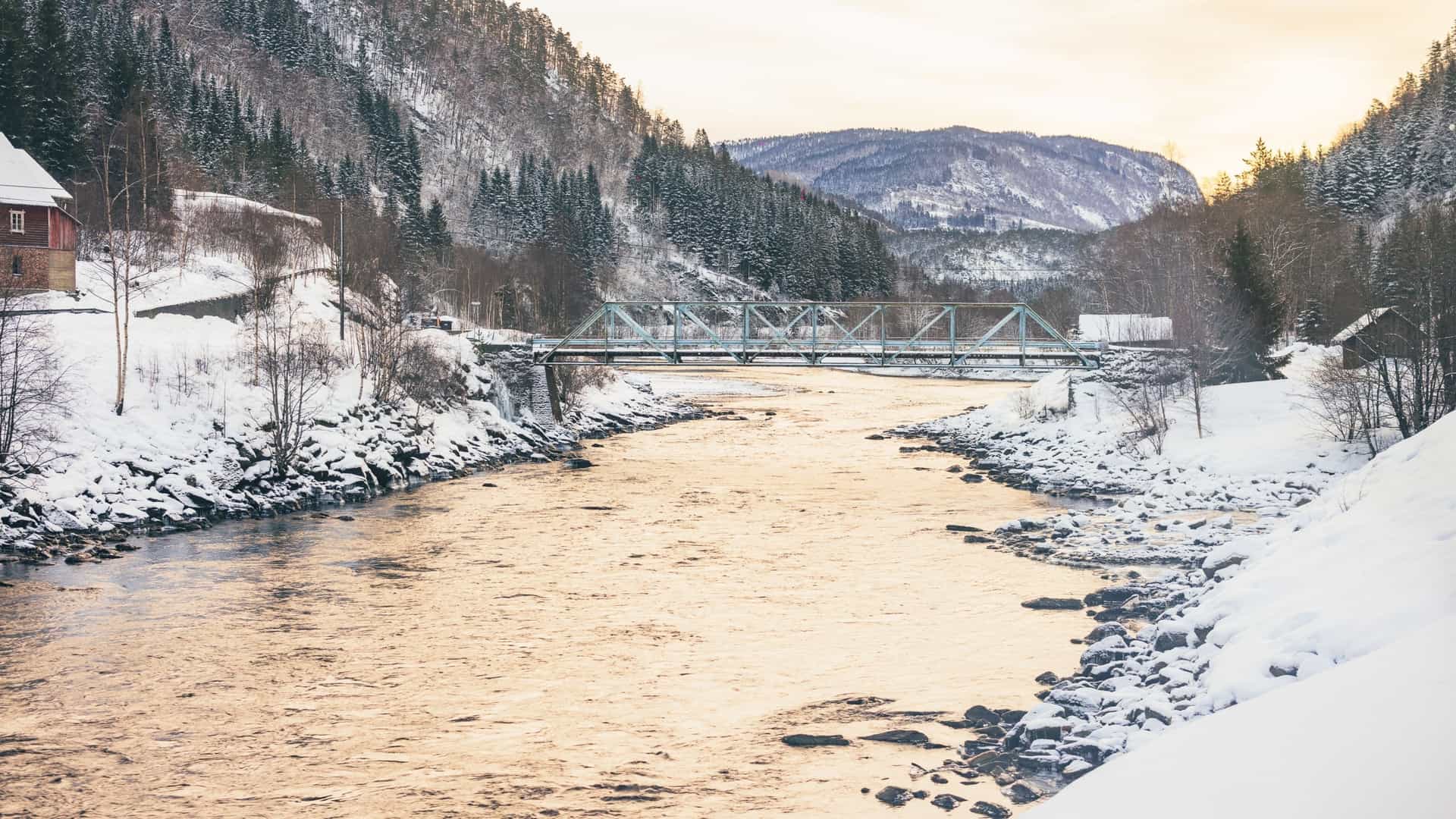 A snow-covered winter landscape in Fjordland, Norway, featuring a wide, slow-flowing river and a light blue truss bridge surrounded by evergreen-covered mountains.