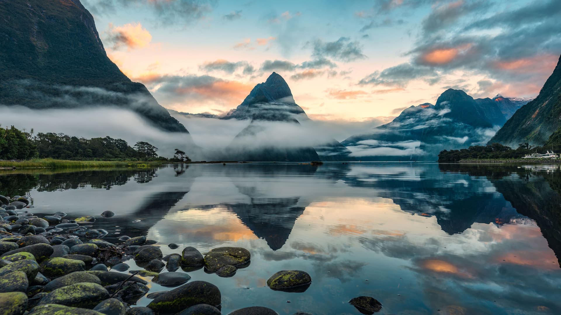 A serene sunrise view of Milford Sound in Fiordland National Park, New Zealand, with the iconic Mitre Peak reflected in the perfectly still water and clouds hugging the surrounding mountains.