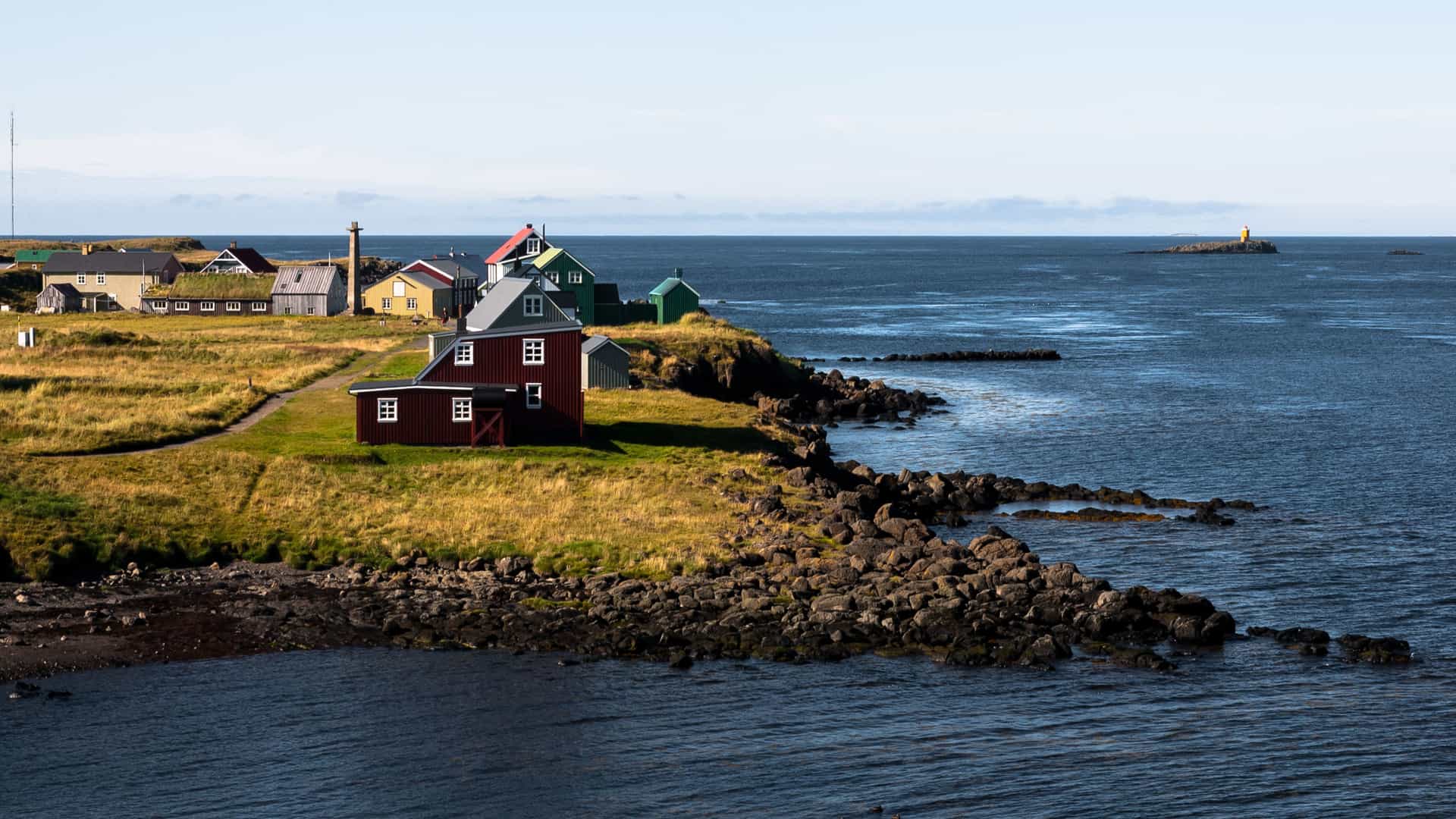 A charming view of colorful houses clustered on the green and rocky shoreline of Flatey Island, Iceland, with a lighthouse and a calm ocean stretching to the horizon.