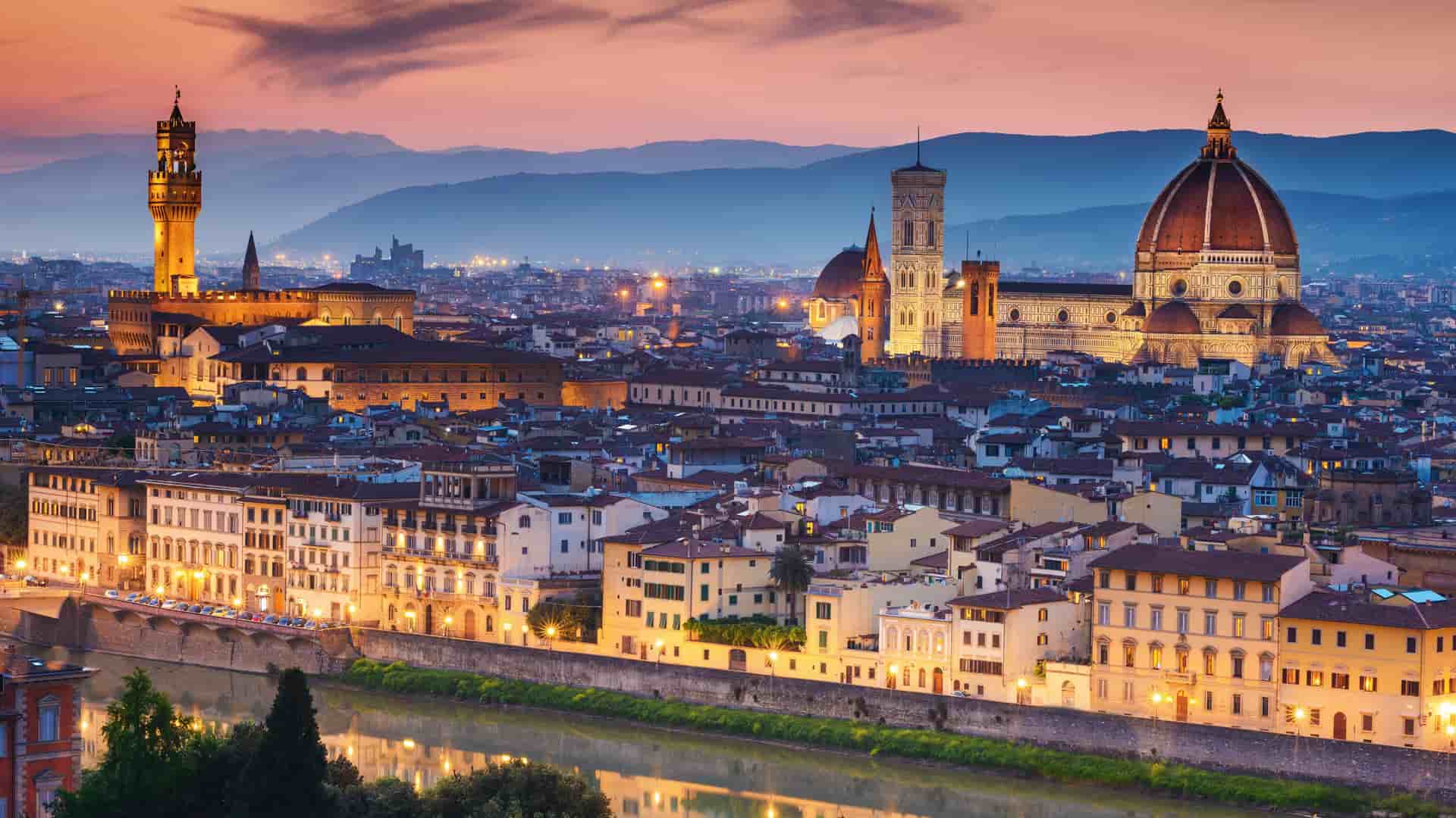 A beautiful twilight cityscape of Florence, Italy, with the iconic Duomo and Ponte Vecchio visible along the Arno River, and the cityscape's warm lights illuminating the historic buildings as the sky turns a vibrant orange.
