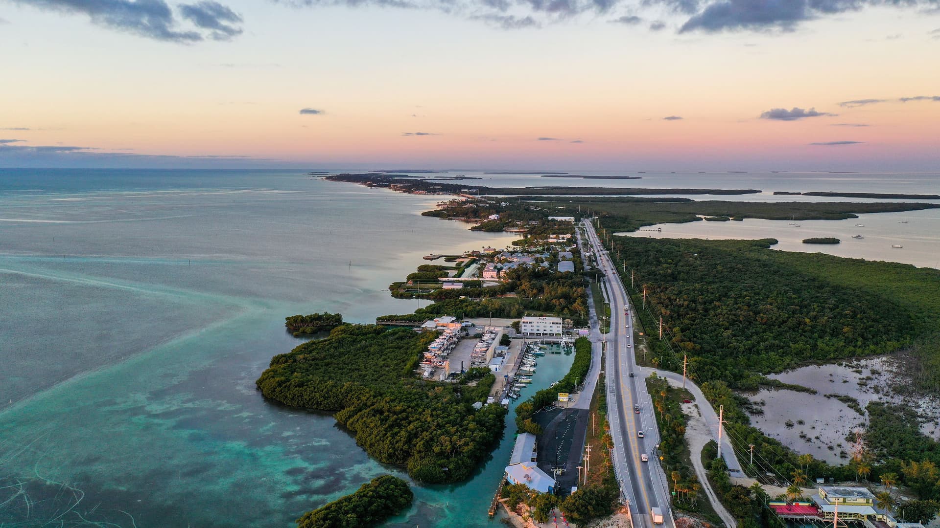 An aerial shot of the Florida Keys at sunset, showing a long highway stretching along the thin strip of land, with the calm, turquoise Florida Strait on one side and a lush mangrove coastline on the other.