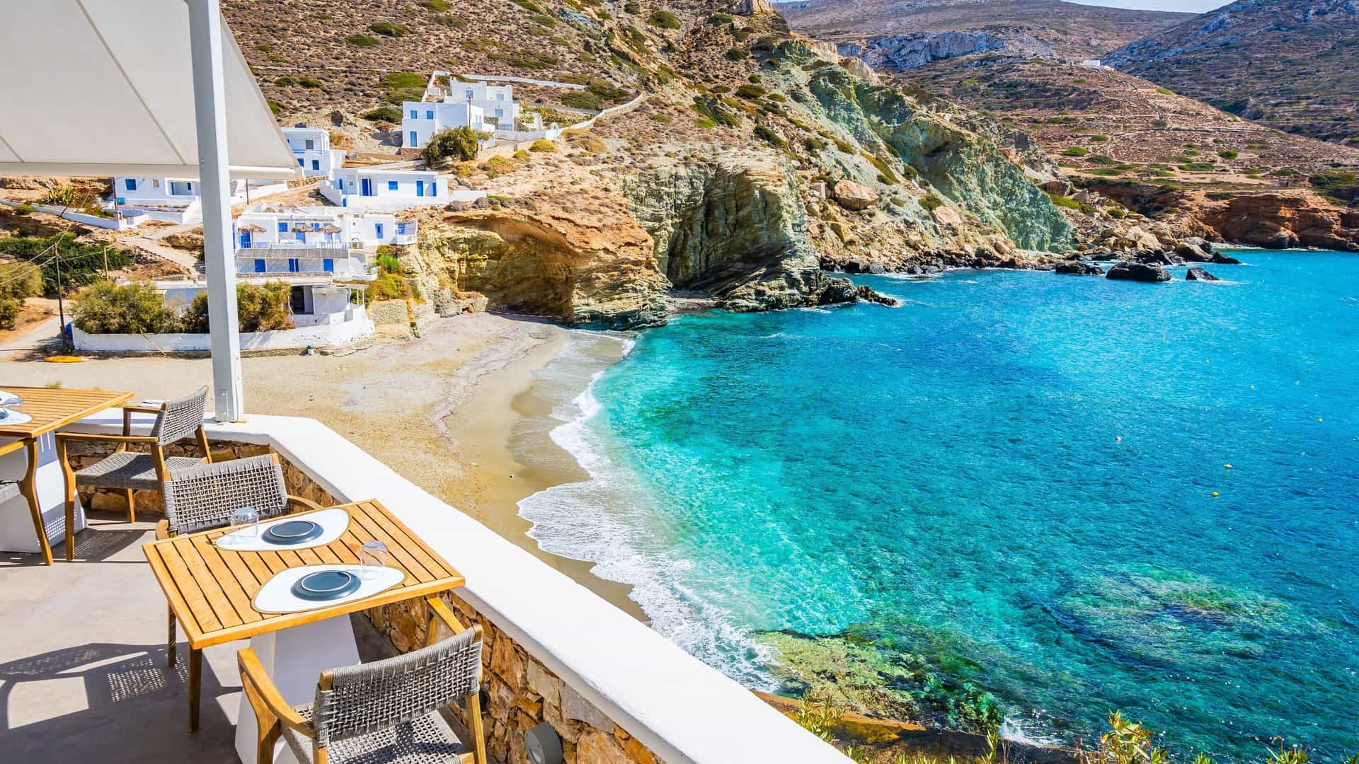 A high-angle view from a restaurant patio overlooking the stunning turquoise waters of a secluded bay and a small sandy beach on the picturesque island of Folegandros in Greece.