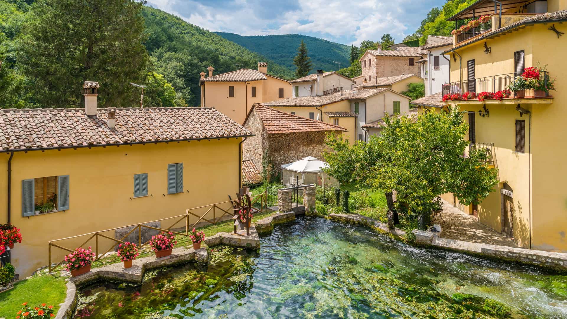 "A tranquil view of Rasiglia, a small village near Foligno, Italy, featuring a crystal-clear stream running through the center of historic stone and stucco houses on a sunny day. "