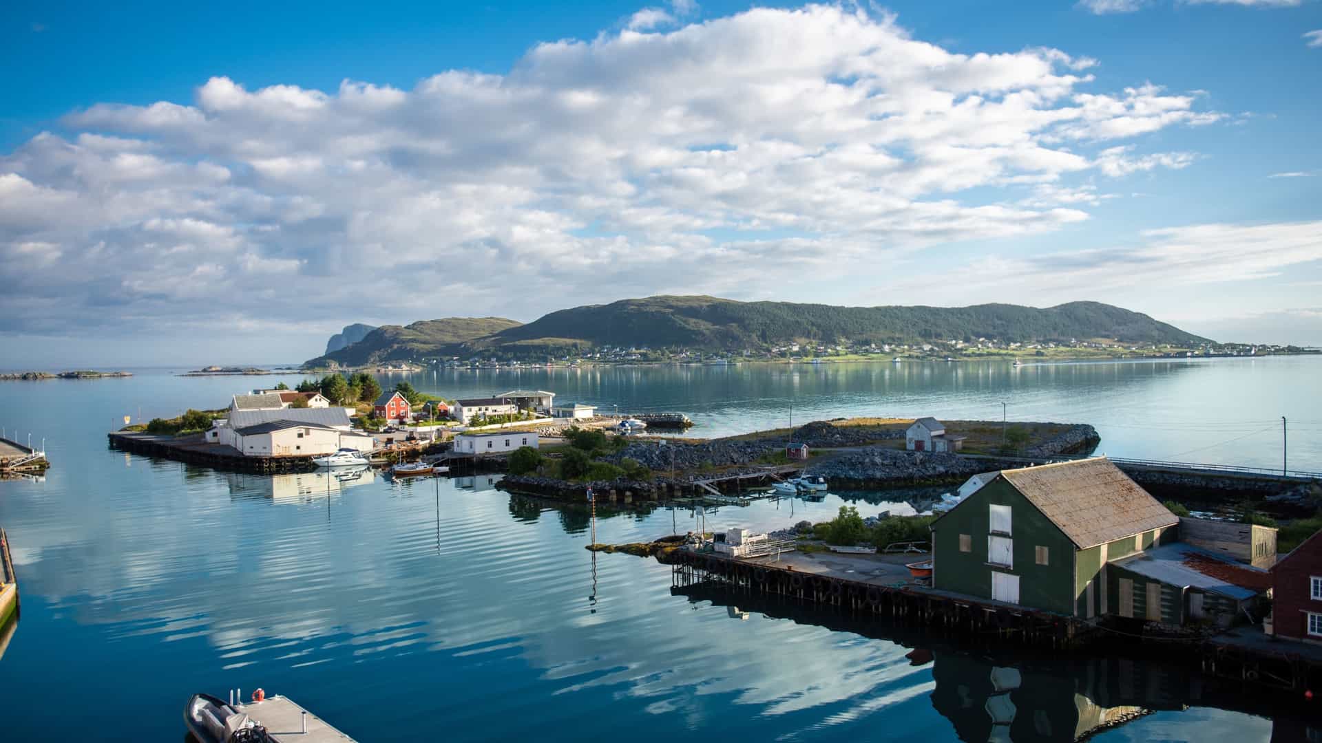 Small, white and green waterfront buildings on an islet in the harbor of Fosnavåg, Norway, under a blue sky.