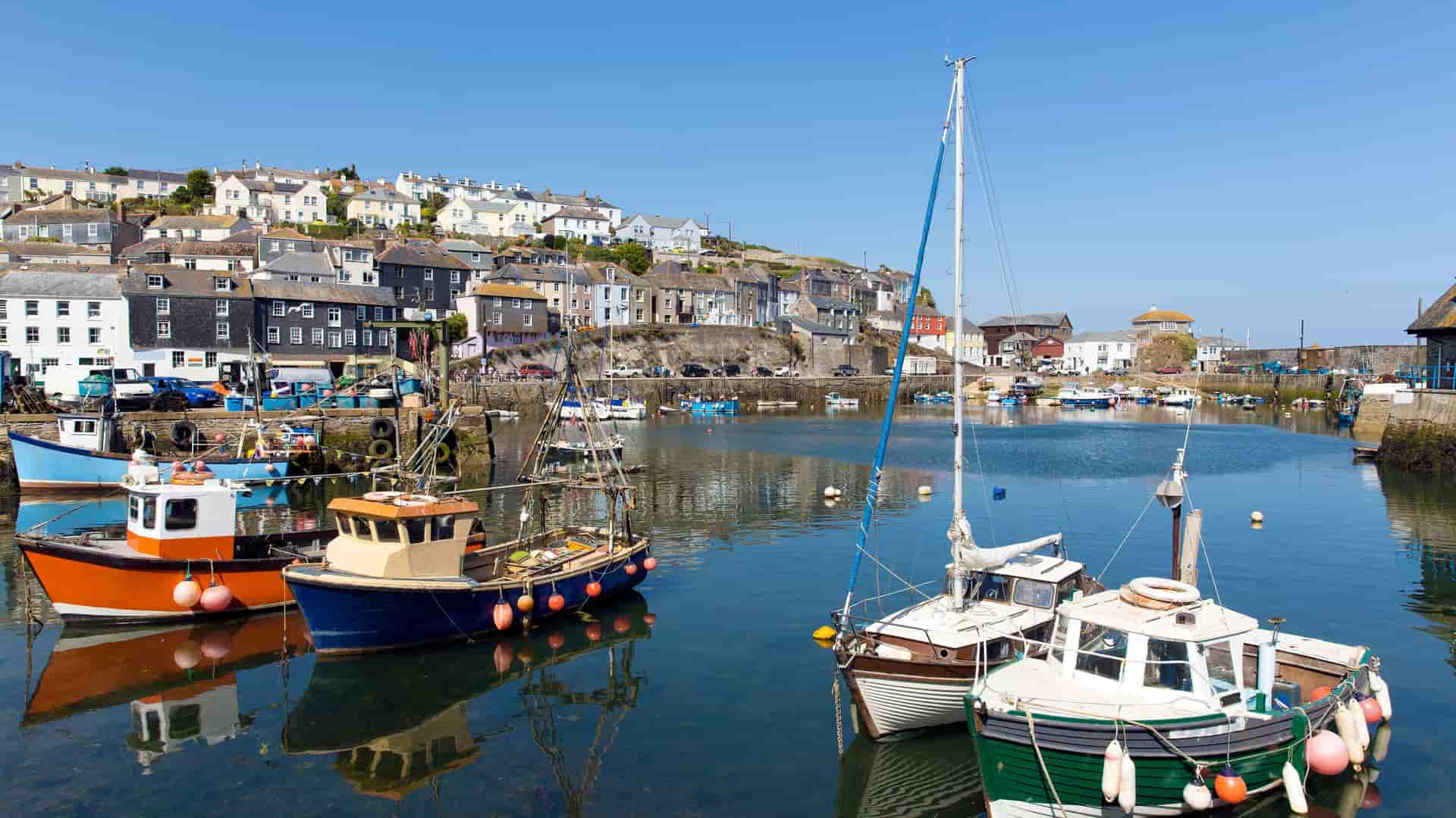 A bright, sunlit harbor scene in Fowey, Cornwall, featuring colorful fishing boats and yachts moored in the calm water with the town's buildings rising up the hill in the background.