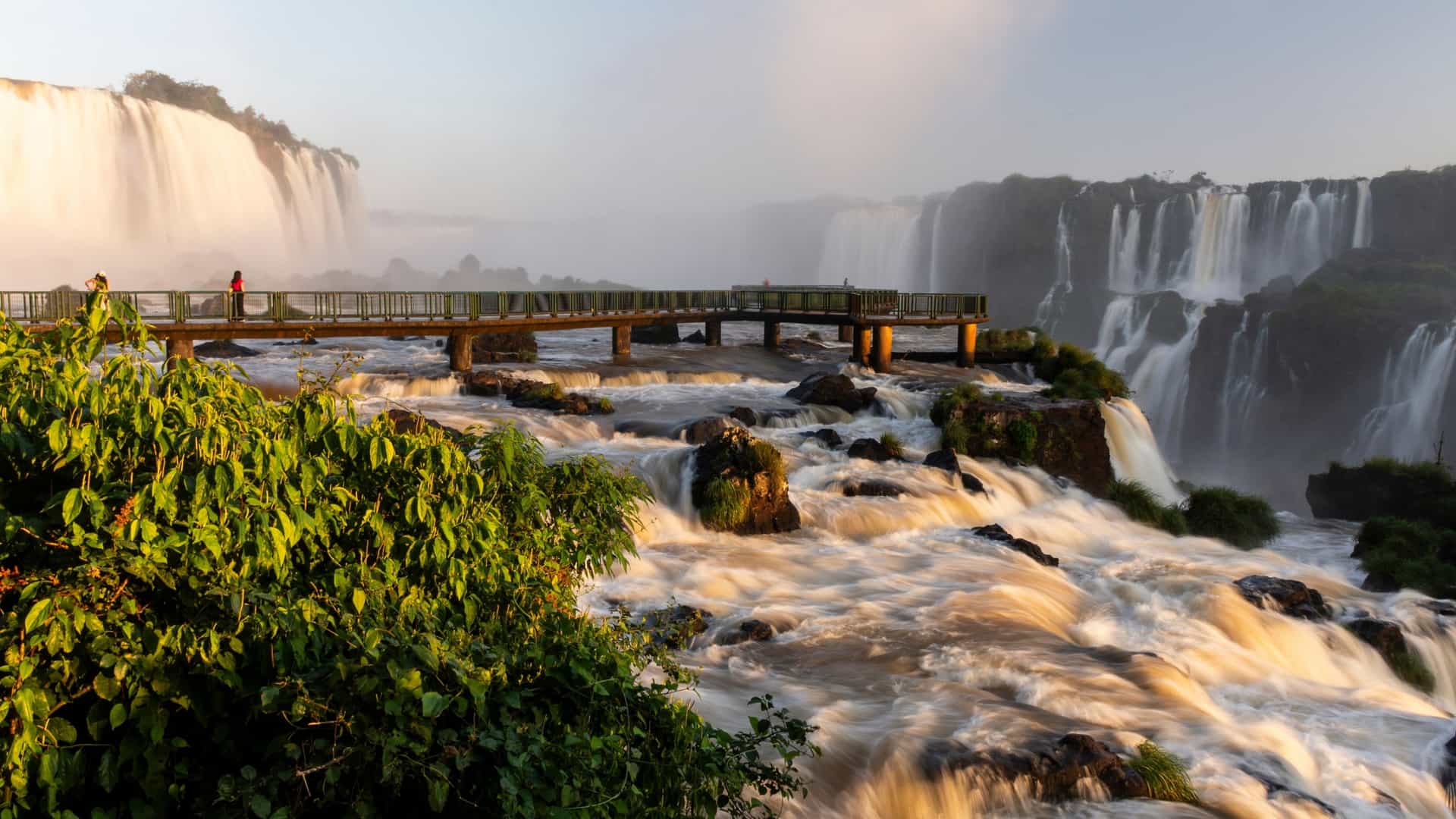 Tourists on a viewing bridge near the powerful, misty cascades of Iguazu Falls (Foz do Iguaçu) on the border between Brazil and Argentina, seen at sunrise.