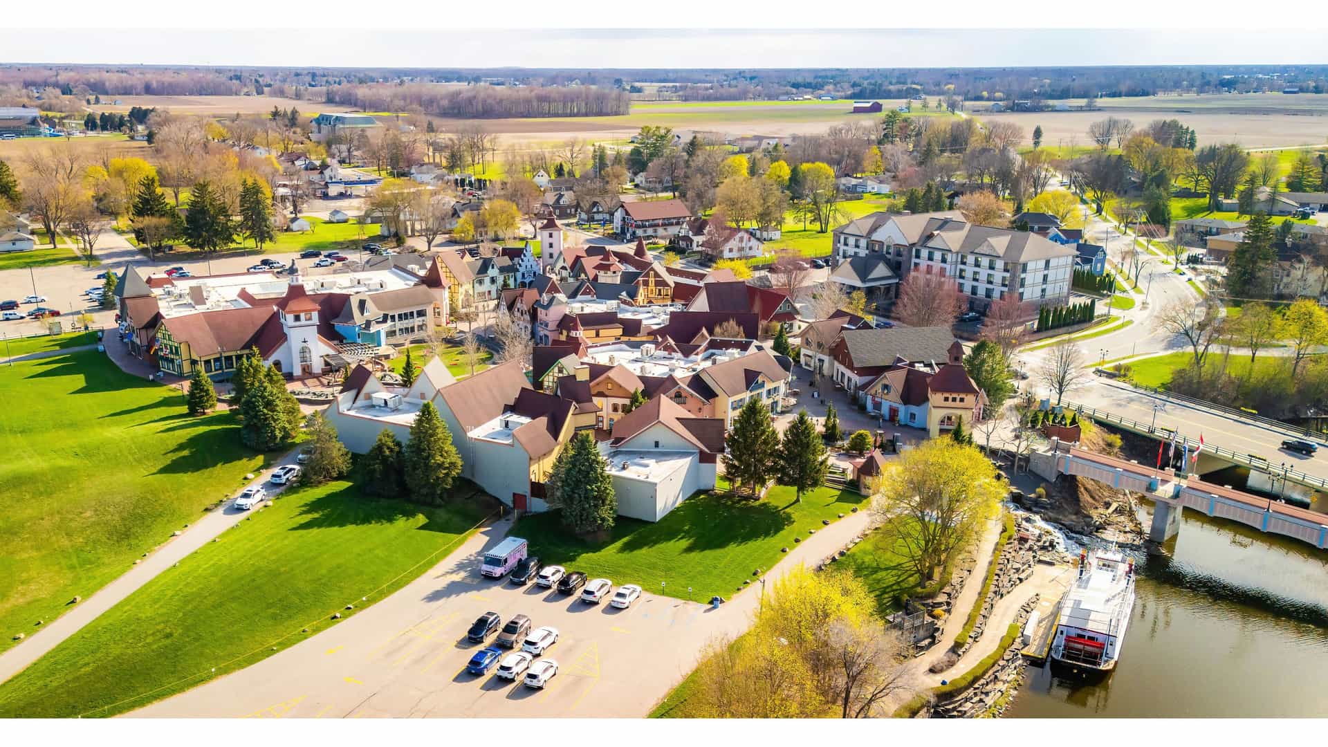 "An aerial view of the Bavarian-style town of Frankenmuth, Michigan, showing its unique architecture, a riverboat on the Cass River, and surrounding green fields. "