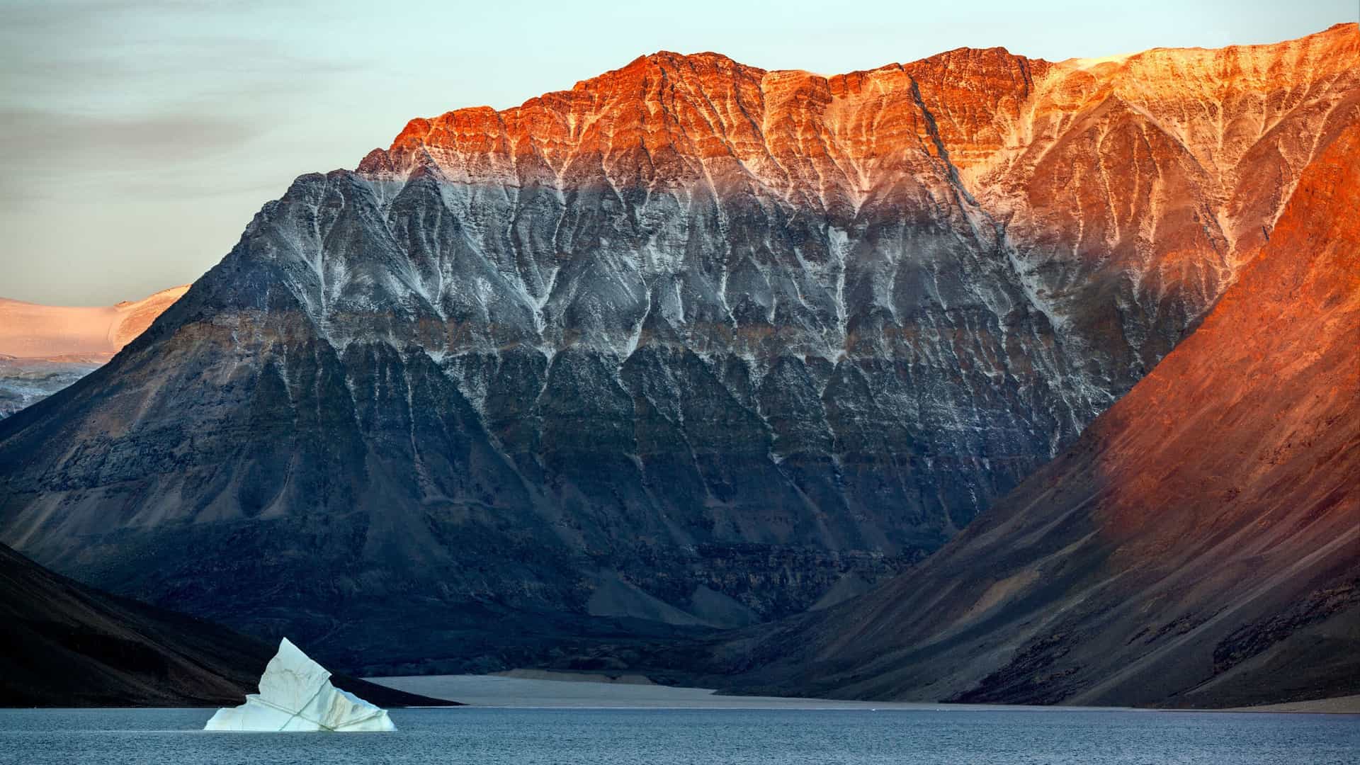 A majestic Arctic landscape with a massive, striped mountain illuminated by the setting sun, towering over the calm waters of the Franz Josef Fjord in Greenland, with a large iceberg floating in the foreground.