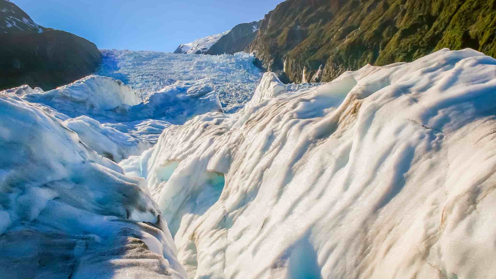"A stunning close-up of the icy, snow-covered surface of the Franz Josef Glacier in New Zealand, showing the deep crevasses and jagged ice formations leading up the mountainside. "