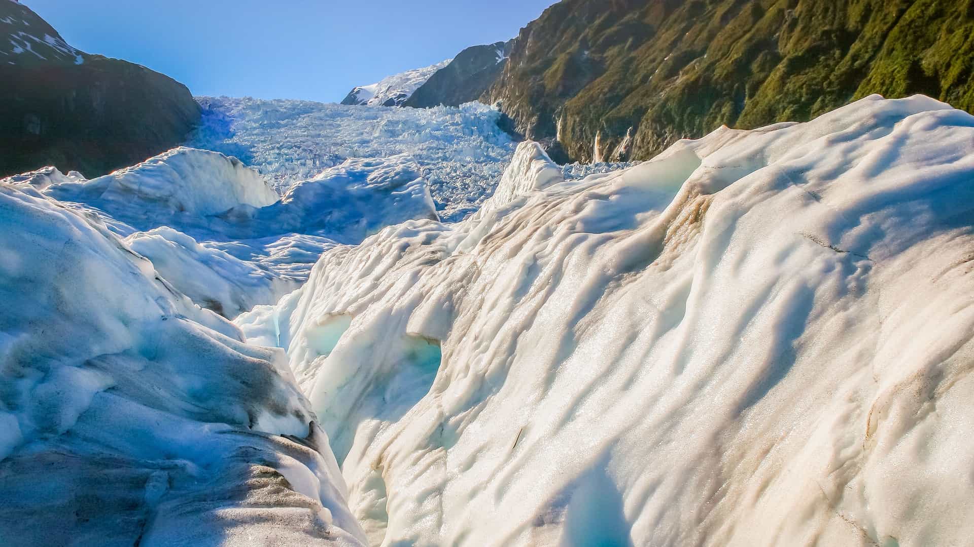"A stunning close-up of the icy, snow-covered surface of the Franz Josef Glacier in New Zealand, showing the deep crevasses and jagged ice formations leading up the mountainside. "