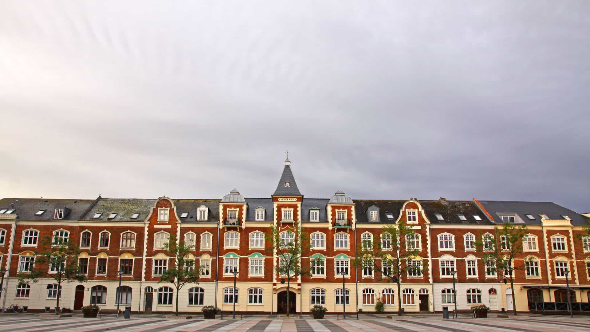 A long, symmetrical brick building with a central spire and white window frames, featuring a row of trees in front on a cobblestone plaza in the city of Fredericia, Denmark.