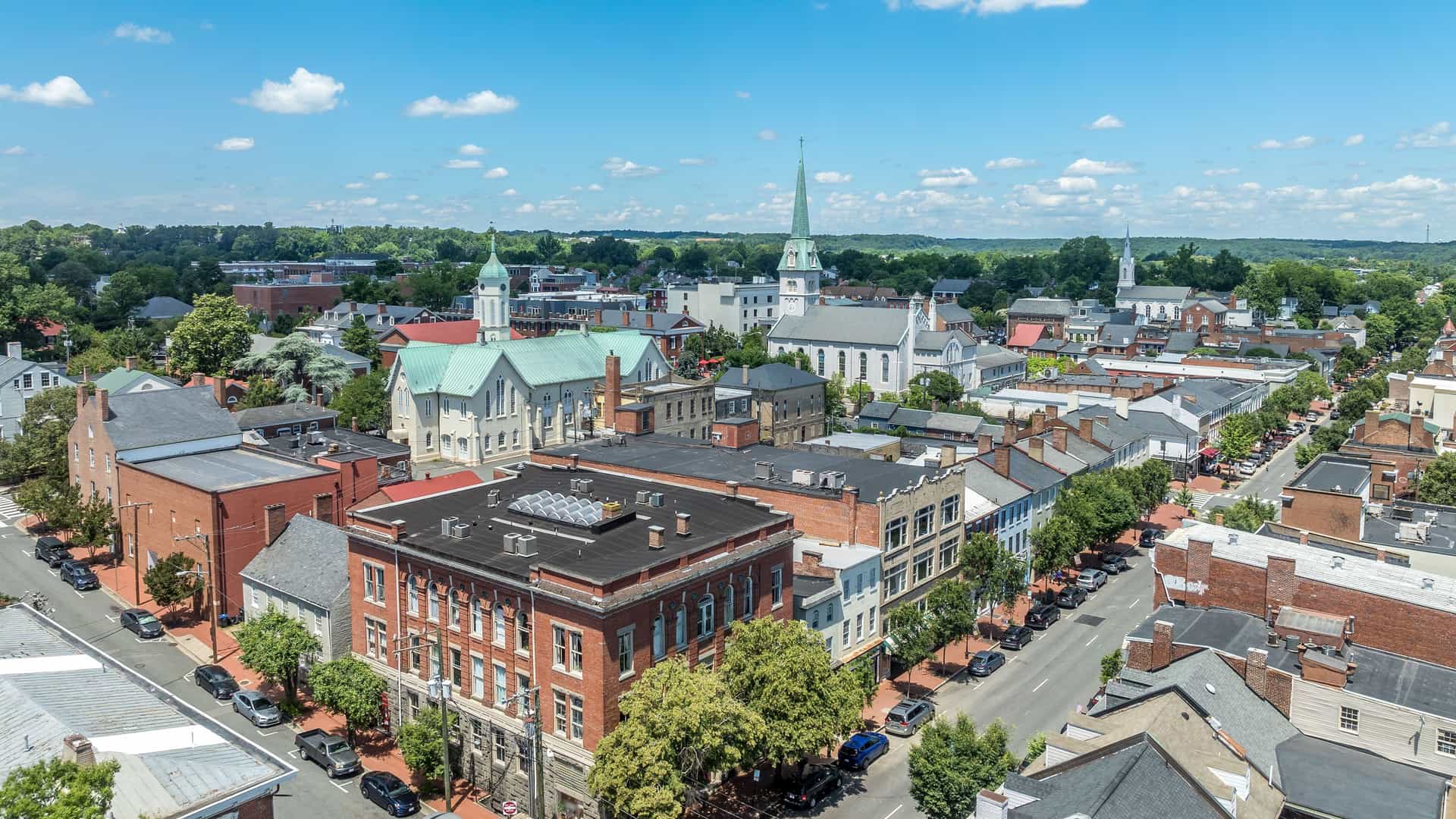 "An aerial view of the historic downtown area of Fredericksburg, Virginia, with its blend of red brick and colonial-style buildings and prominent church steeples under a bright blue sky. "