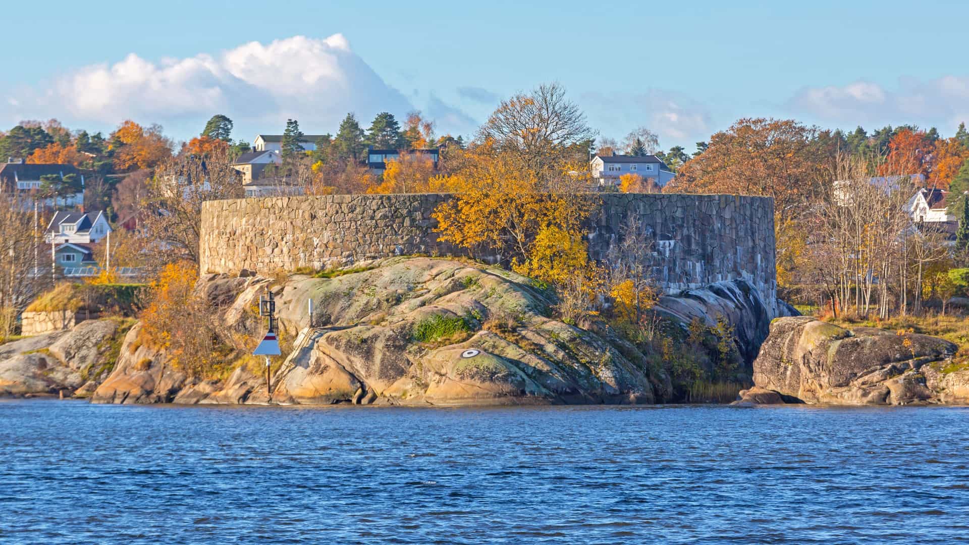 A historic, round stone fortress on a rocky outcrop along the water in Fredrikstad, Norway, with autumn trees and residential houses on the hill in the background.