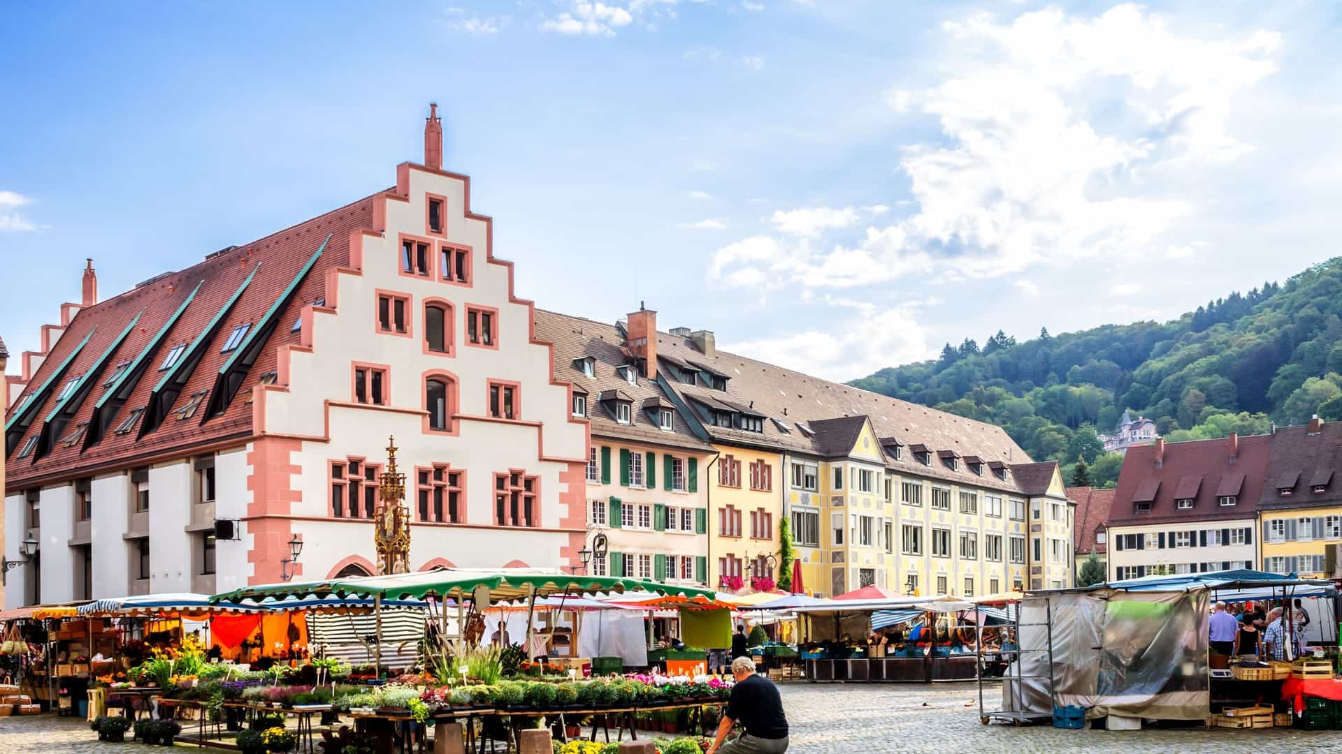 A wide-angle shot of the bustling farmers market in the center of Freiburg, Germany, with colorful stalls and the historic, gabled facades of the old city in the background.