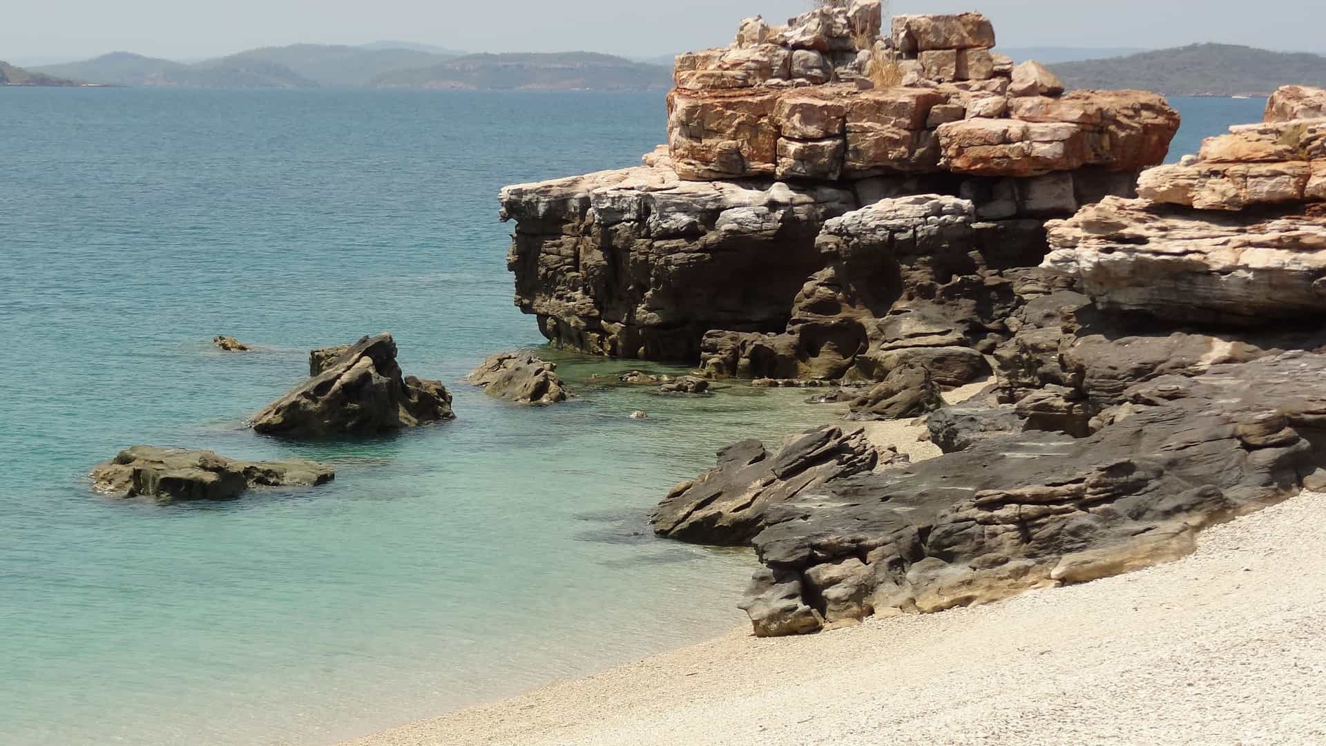 A close-up view of the rugged, layered rock formations and sandy shore of Freshwater Cove, Australia, with a tranquil turquoise ocean and distant hills under a clear sky.