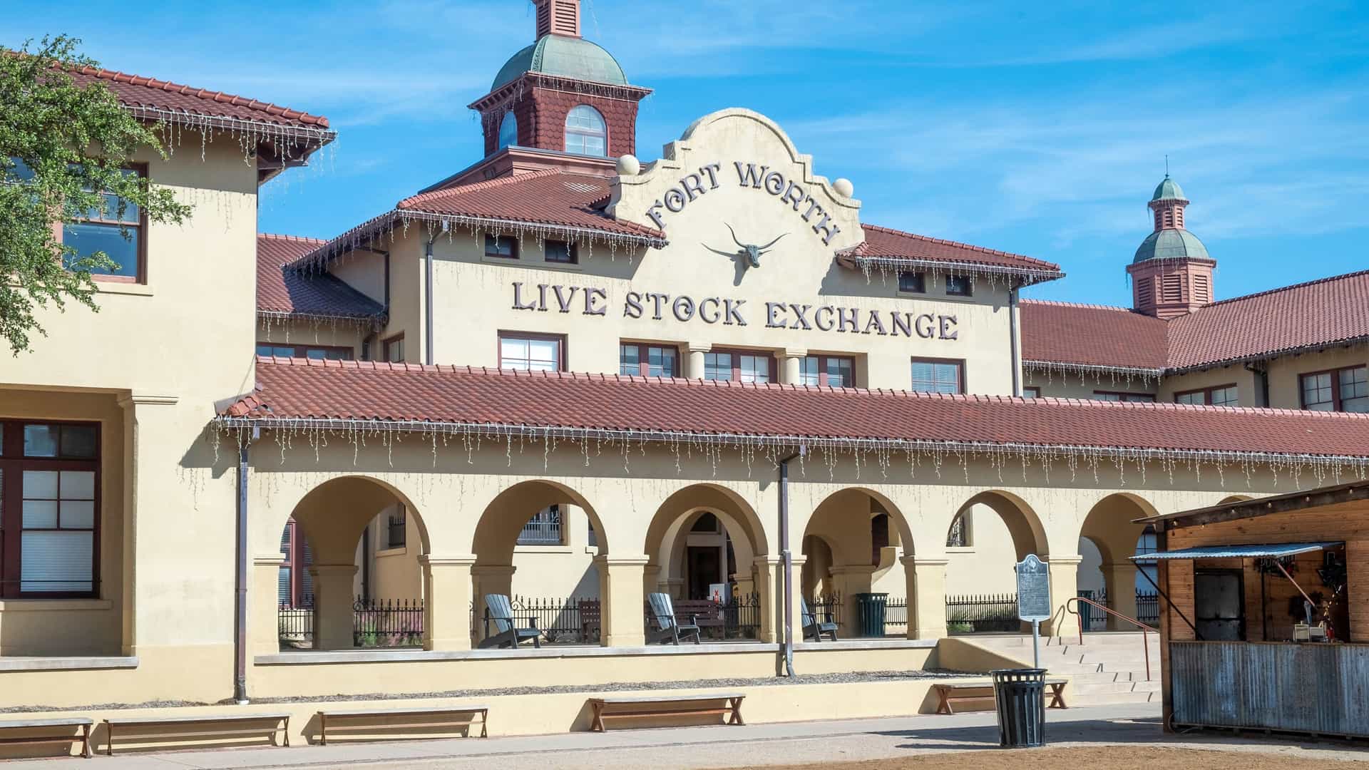 "A classic facade of the historic Fort Worth Live Stock Exchange in Texas, a landmark with arched entryways and a sign featuring a longhorn emblem under a bright blue sky. "