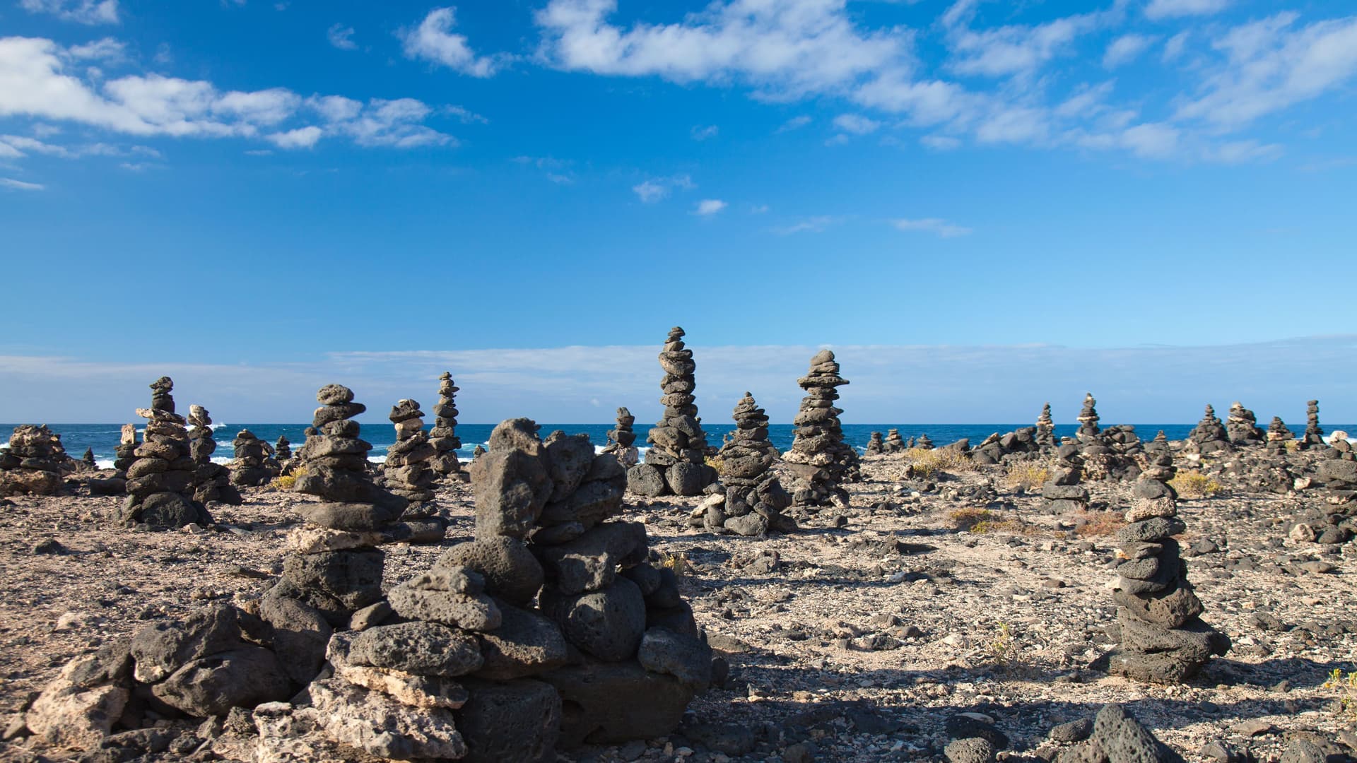 A panoramic view of the rocky landscape of Fuerteventura, Spain, showing numerous cairns (stacked stones) against a background of the blue Atlantic Ocean and a partly cloudy sky.